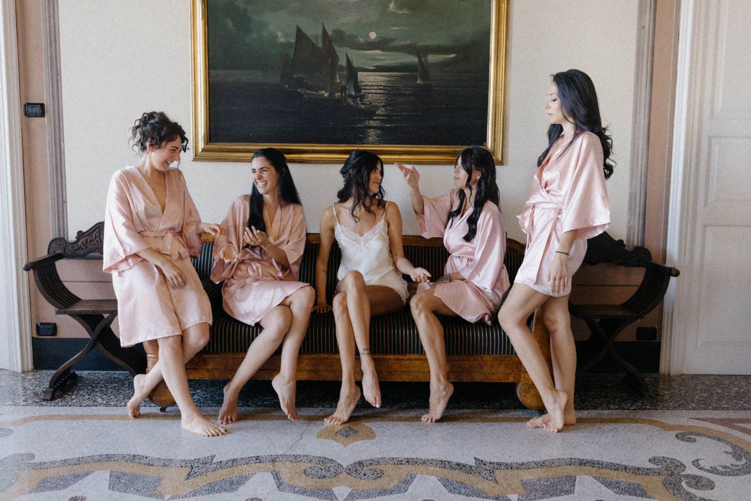 A group of people in pink robes sit together on a wooden floor during a wedding preparation event.