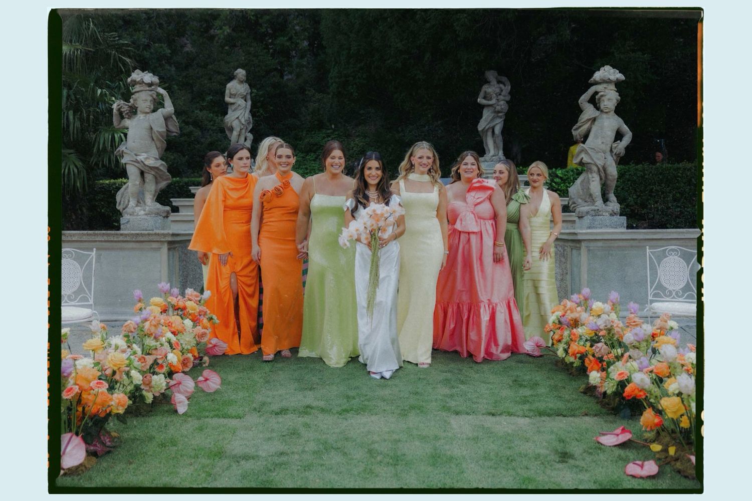 Wedding party members pose on grass with colorful floral arrangements and coordinated orange and pink dresses.