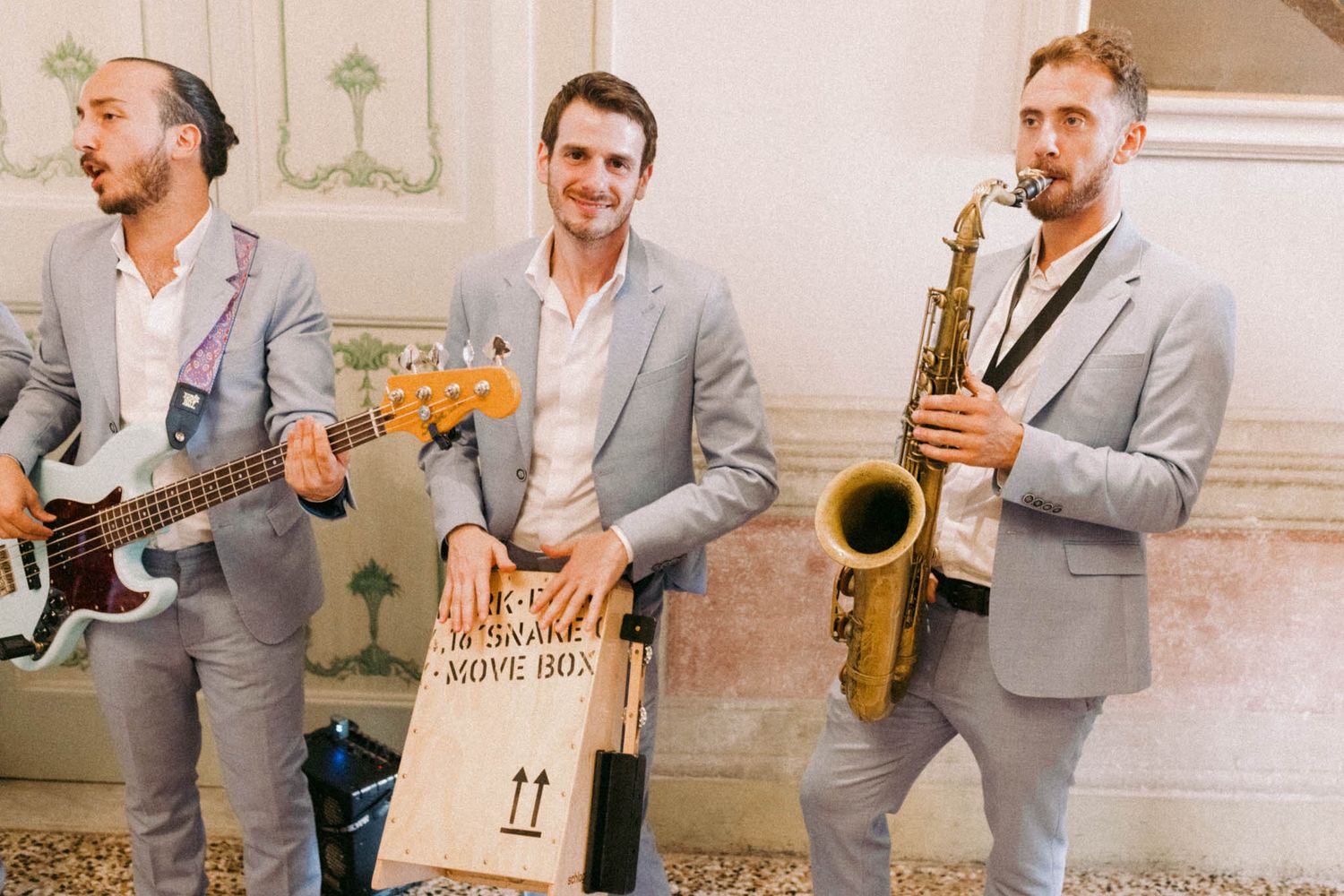 A group in matching gray suits plays musical instruments while standing in a line against a light-colored wall.