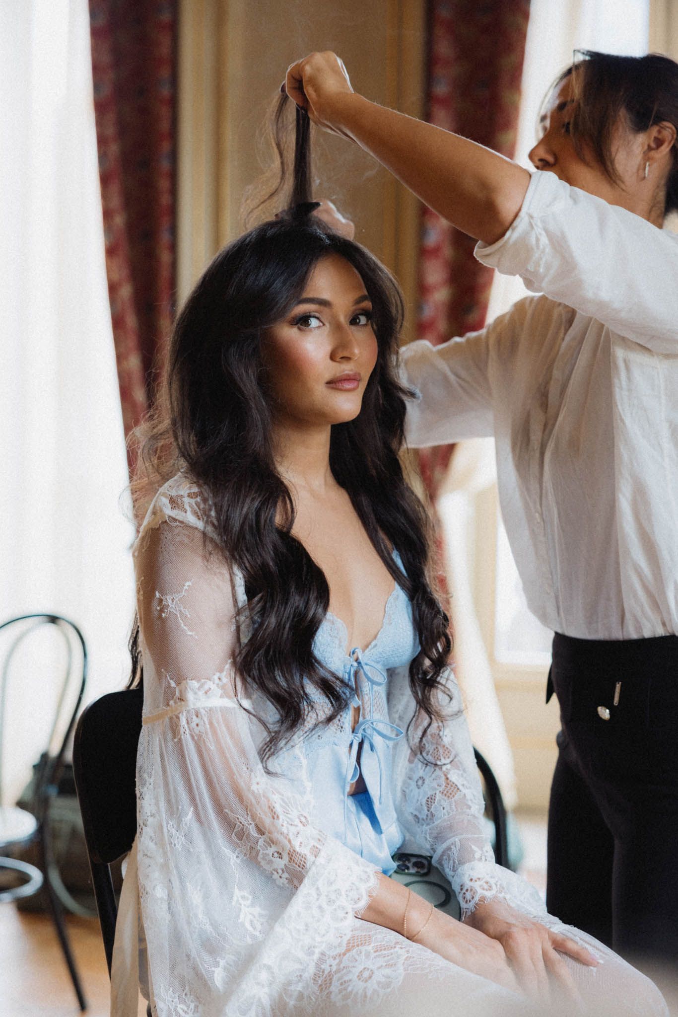 Romantic wedding preparation scene with couple in white lace robe and formal attire by window with soft lighting.