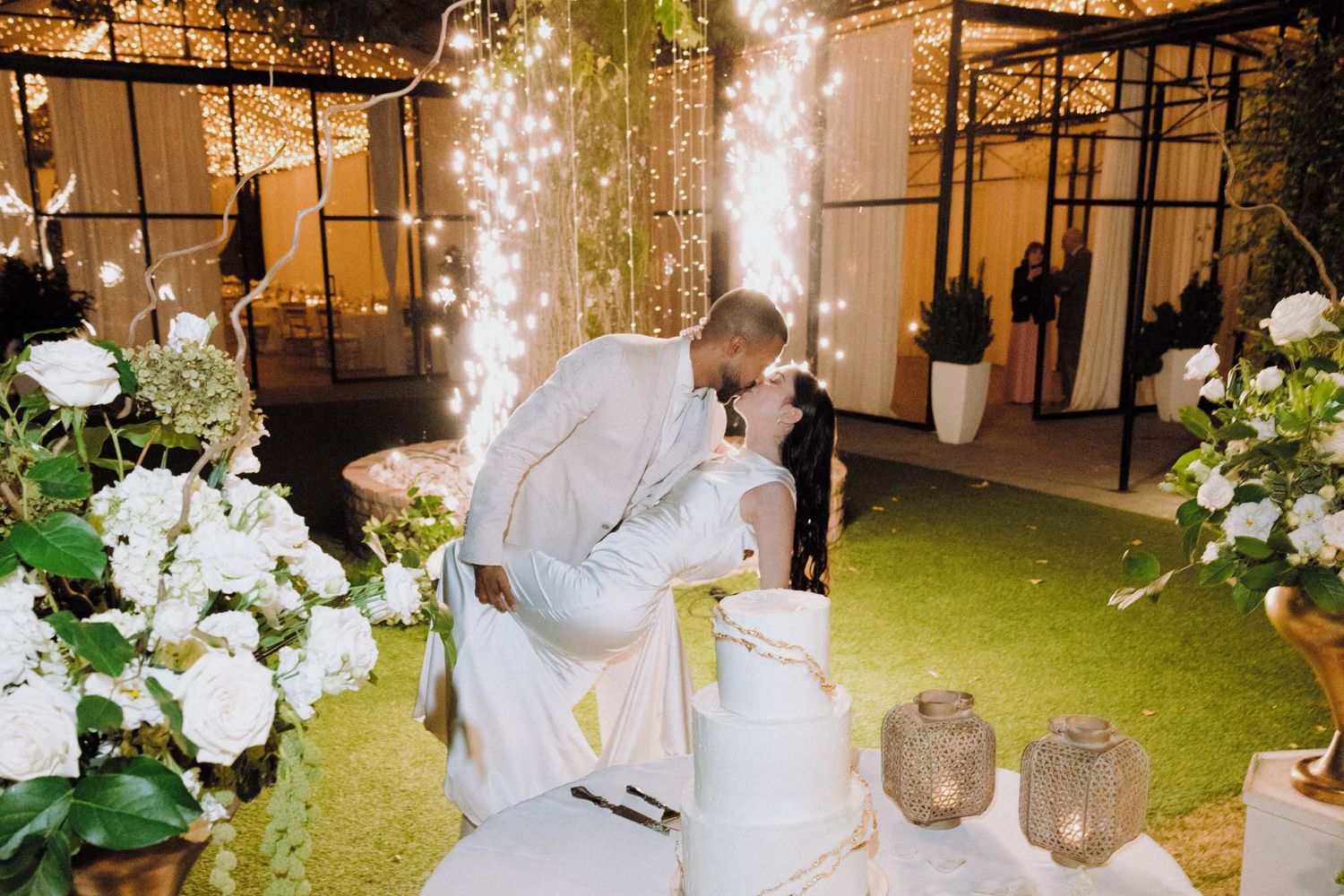 A romantic wedding moment captured under twinkling lights and white floral decorations in an elegant indoor venue.