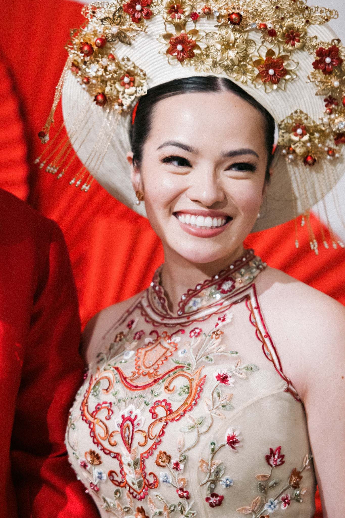 Traditional Chinese bride wearing ornate gold headdress and embroidered qipao dress against red backdrop.