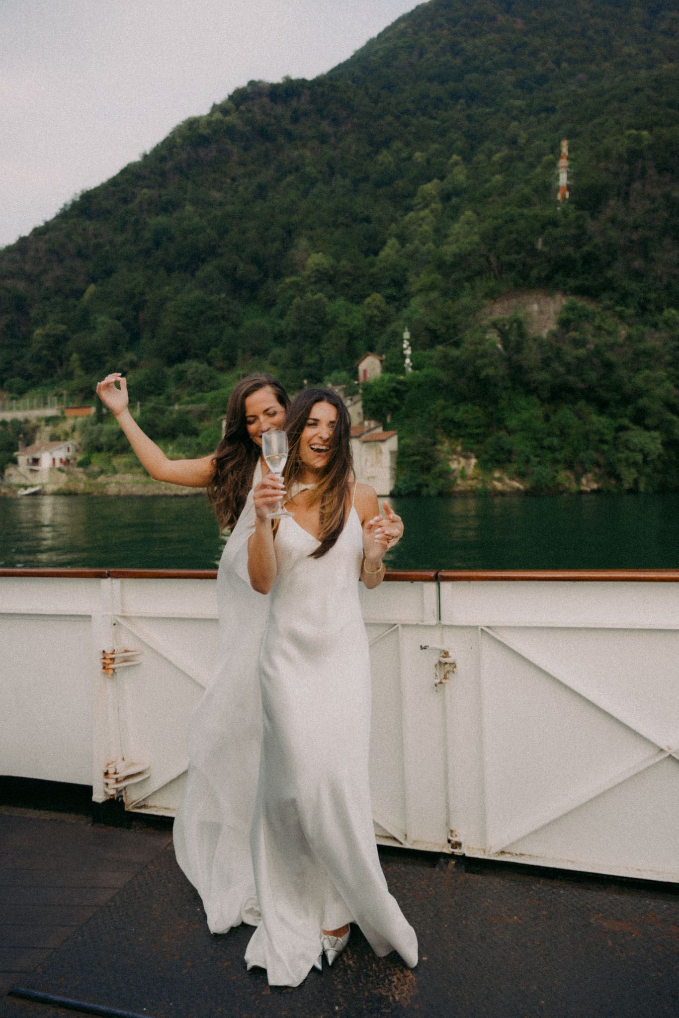 Two people celebrating on a boat deck with champagne glasses against a scenic mountain lake backdrop.