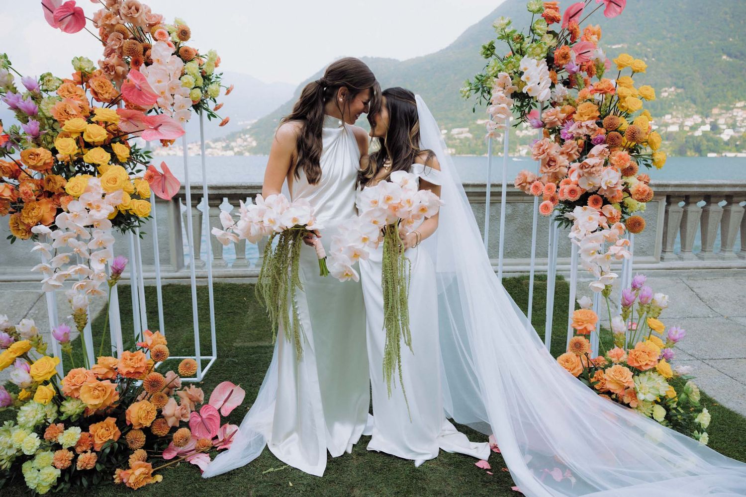 A bride in a flowing white dress poses with vibrant coral and pink floral arrangements by a lakeside terrace.