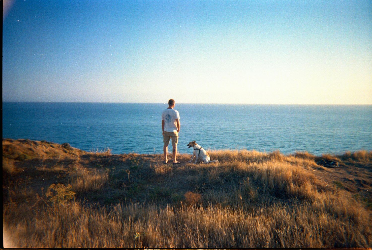 Panoramic sequence showing two figures walking along a coastal cliff path against a blue ocean horizon at sunset.