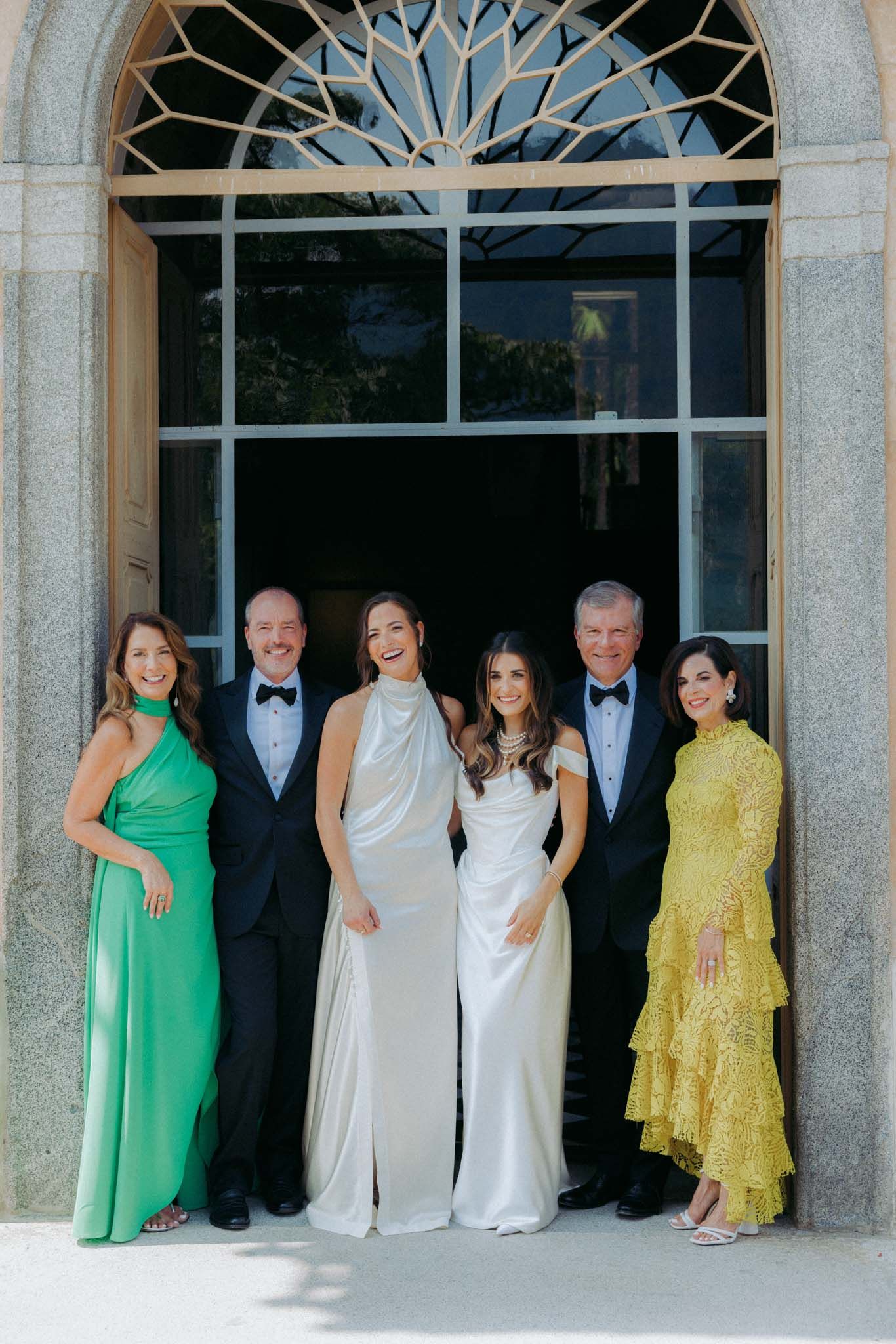 Wedding party members in bright green, yellow, and white dresses pose together under a columned portico.