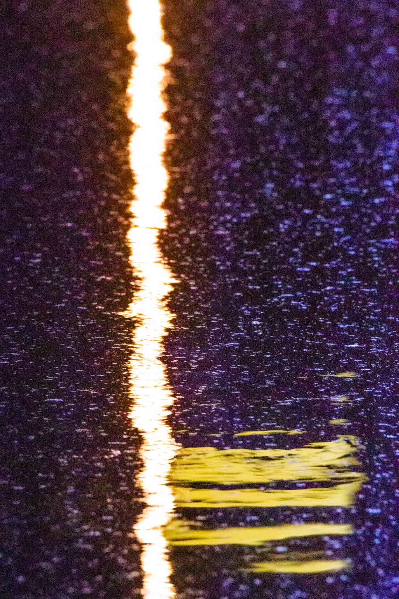 A vertical streak of golden light reflects on wet pavement with purple sparkles in the dark night.