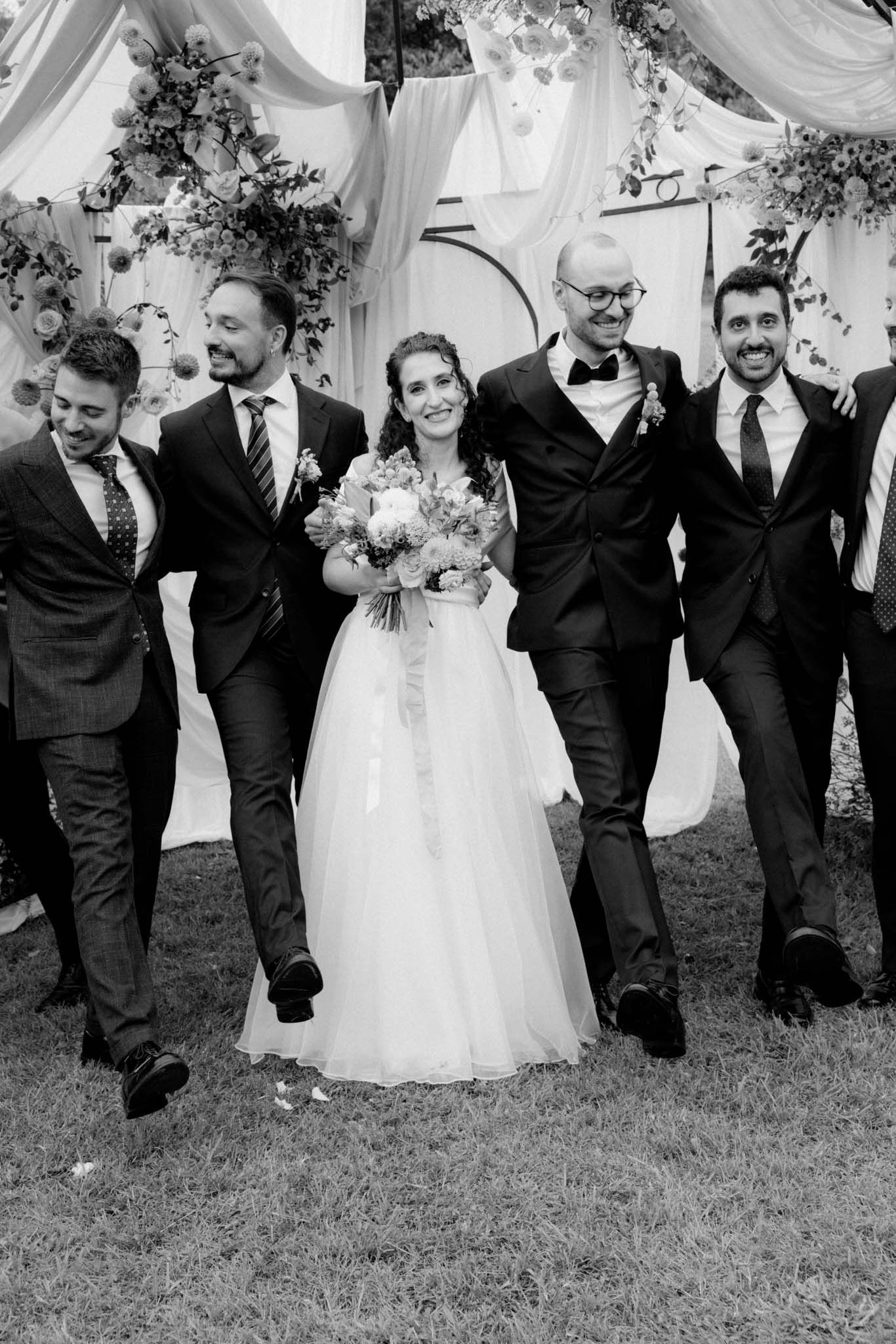 Wedding party in black suits and white dress walk together on grass in playful candid sequence.
