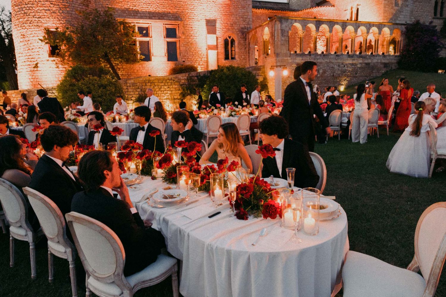 Outdoor evening wedding reception with white tables and warm lighting creating an elegant ambiance in a garden setting.