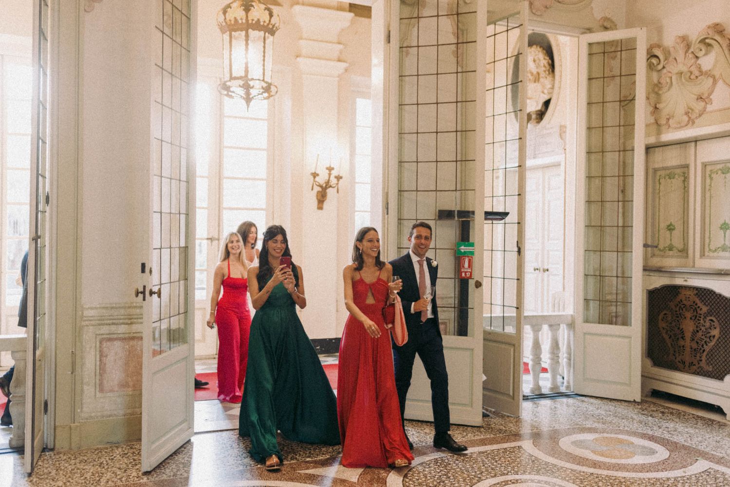 People in formal attire gather in an elegant ballroom with ornate chandeliers and mirrored walls.