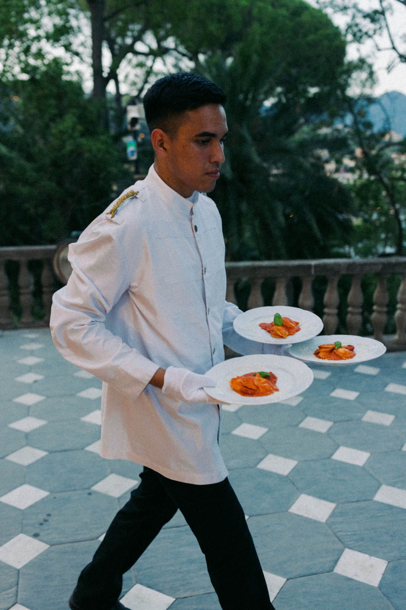 A waiter in a crisp white uniform carries three plates of colorful dishes on an outdoor terrace with geometric floor patterns.