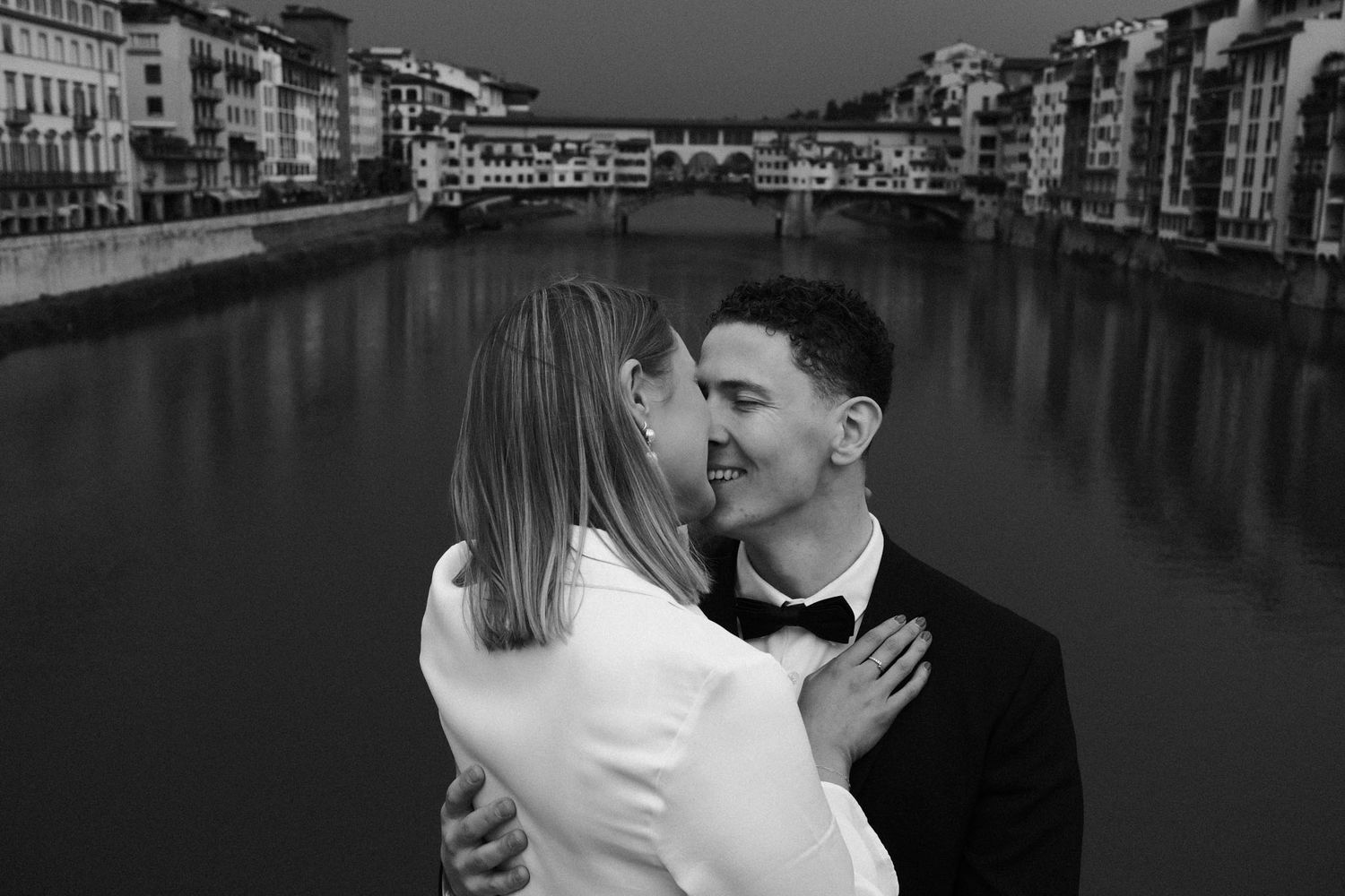 A romantic couple embraces in black and white against the backdrop of Florence's Arno River and historic buildings.