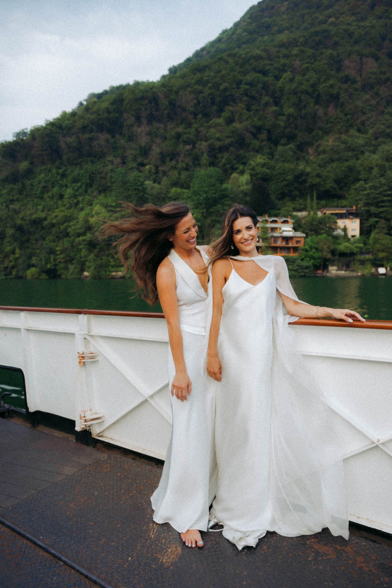 Two people in flowing white dresses dance and twirl together on a boat deck with lush green mountains in the background.