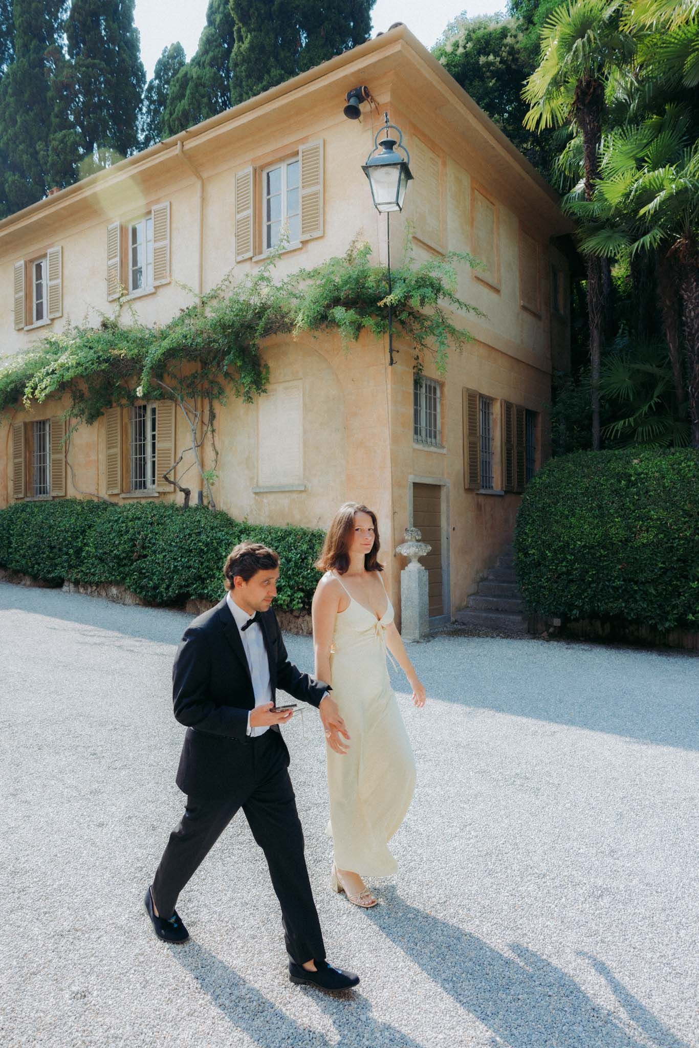 A couple in formal attire walks together on a gravel path in front of a Mediterranean-style villa with climbing vines.