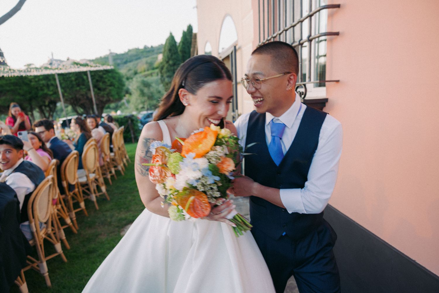 Outdoor wedding ceremony setup with wooden chairs on a lawn facing a pink stucco building at sunset.