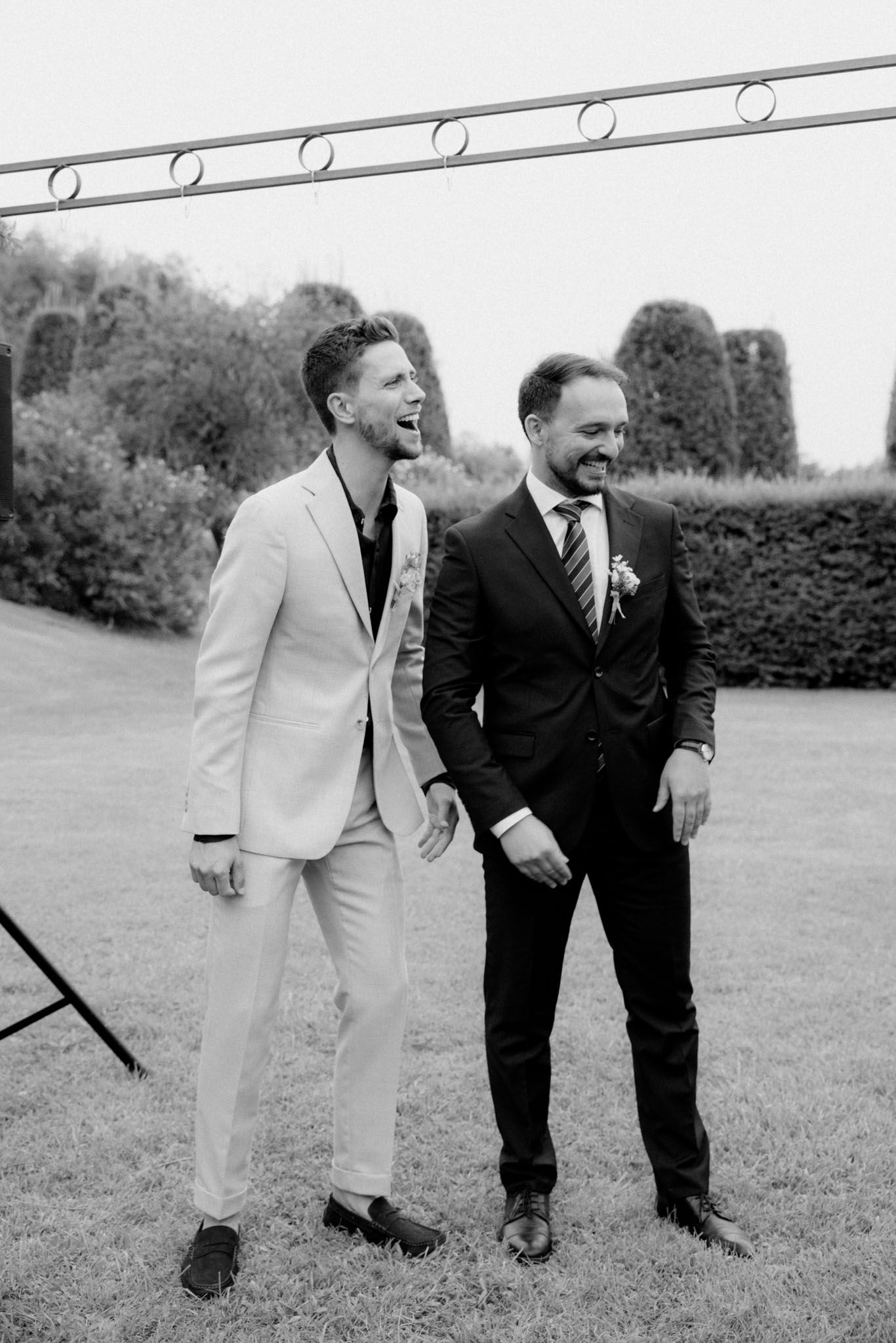 Two people in contrasting black and white suits share a lighthearted moment while looking down at something on the grass.