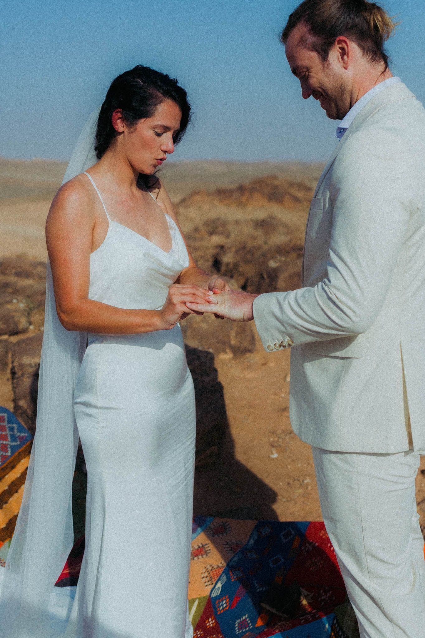 A couple in white attire shares a tender moment against a desert landscape backdrop.