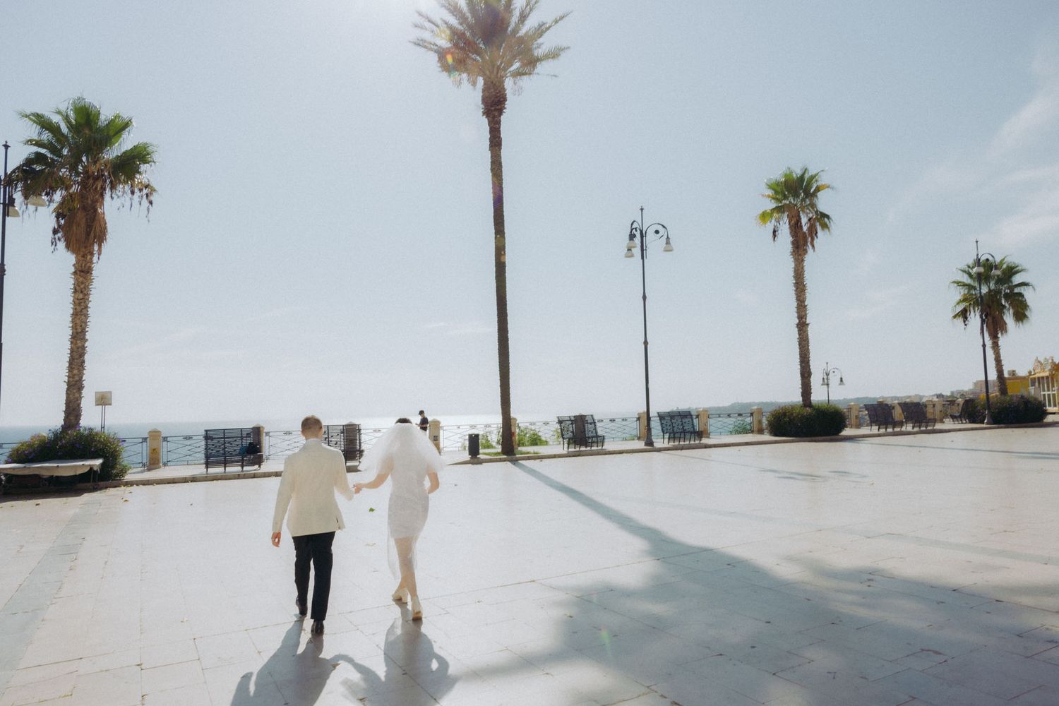 Palm trees line a white concrete walkway against a bright blue sky on a sunny day.