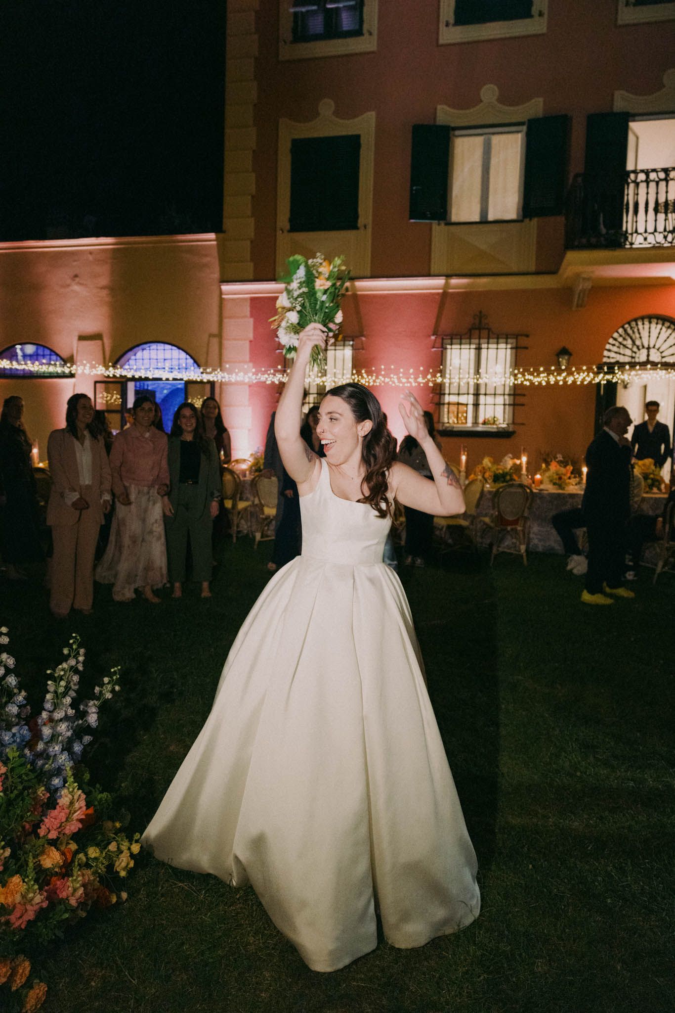 Two people in elegant white wedding gowns dance together at an outdoor nighttime reception with string lights overhead.