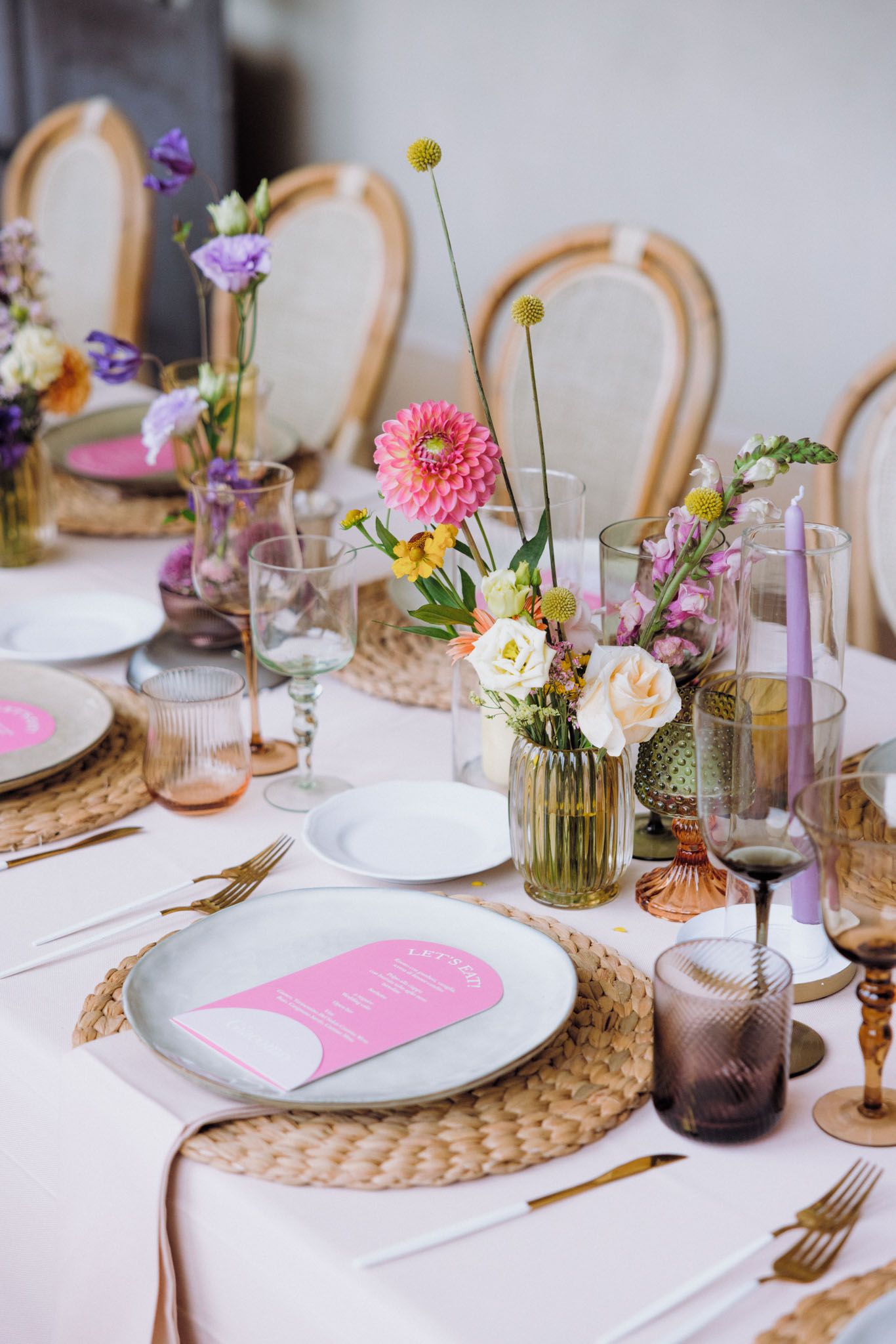 Elegant table setting with pink placemats, woven chargers, white plates, and delicate floral centerpieces in glass vases.