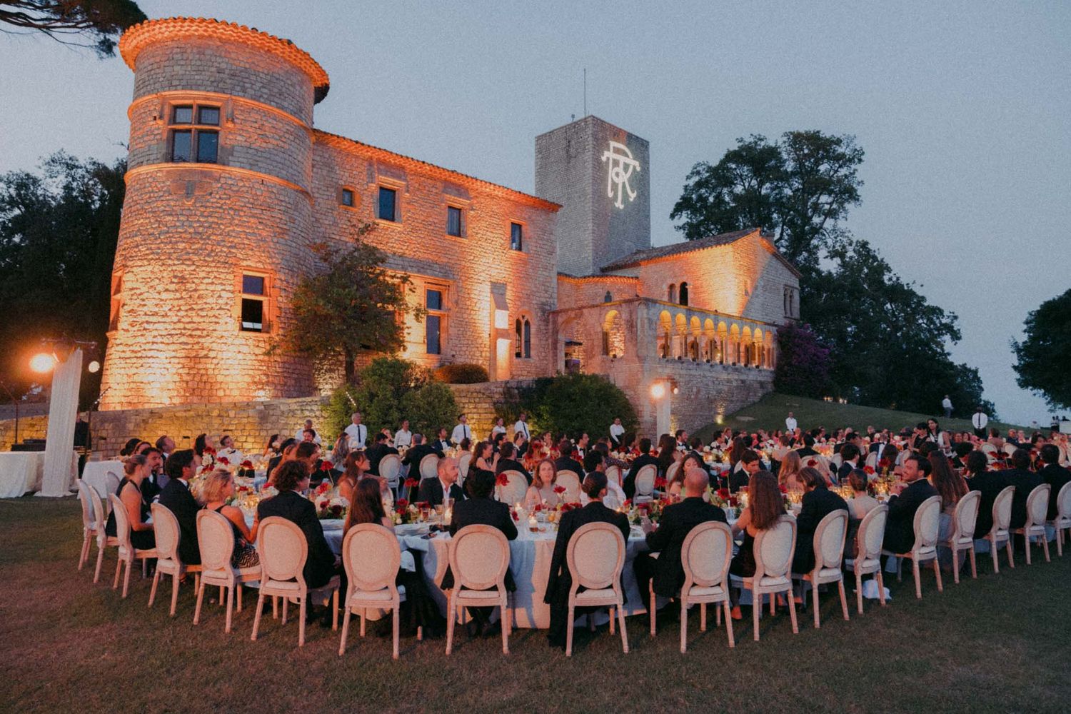 Wedding guests enjoy an outdoor reception dinner at dusk near an illuminated castle-like venue.