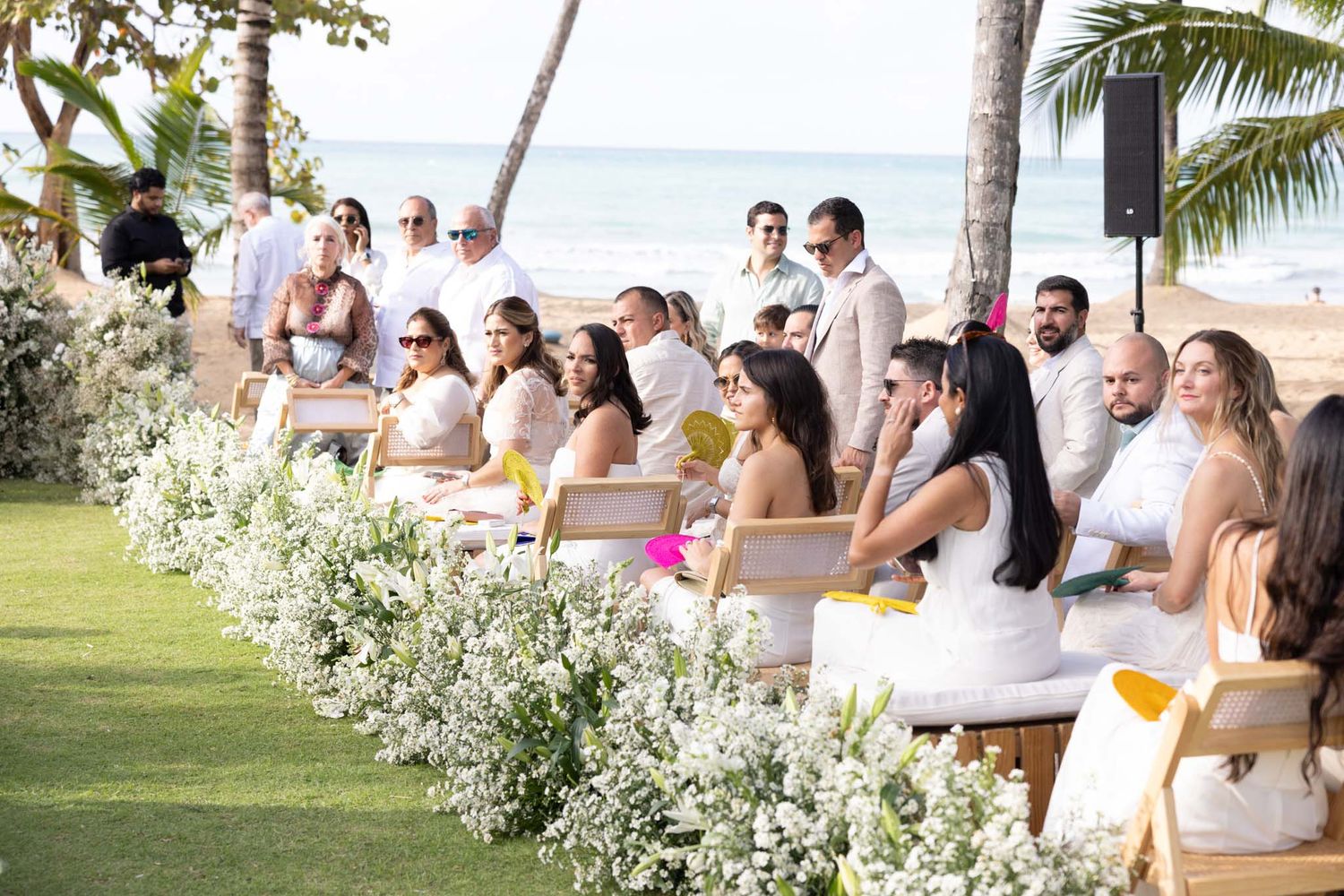 Wedding guests seated in white chairs along a tropical beach path with palm trees and ocean views.