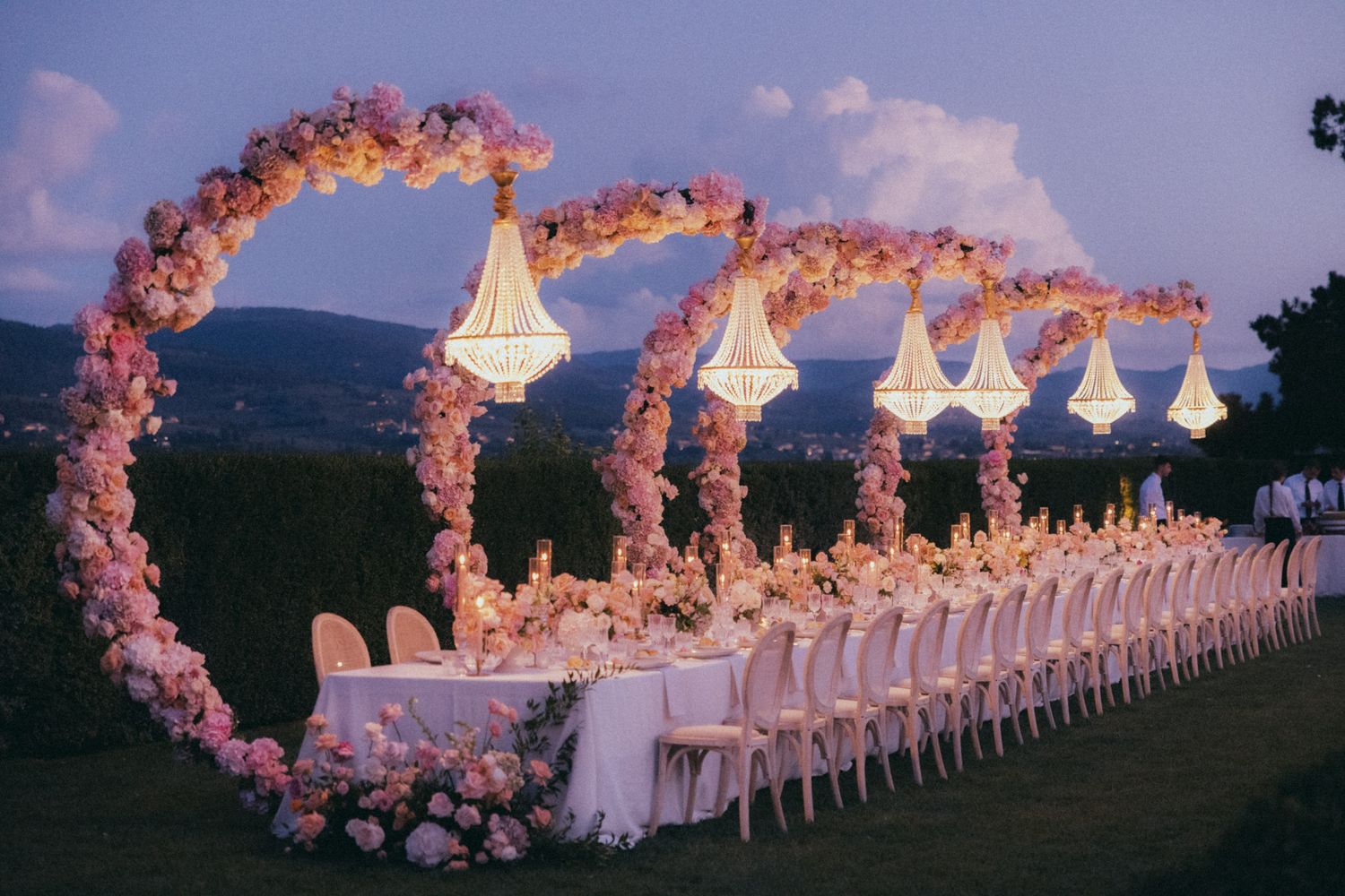 Elegant outdoor wedding reception setup with pink floral arches and white chairs along a scenic mountain view at sunset.