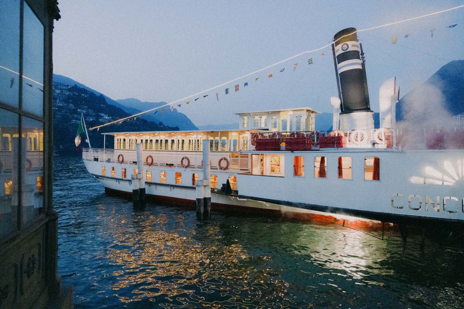 Vintage passenger ferry boats docked at a waterfront terminal with mountains and buildings in the background.