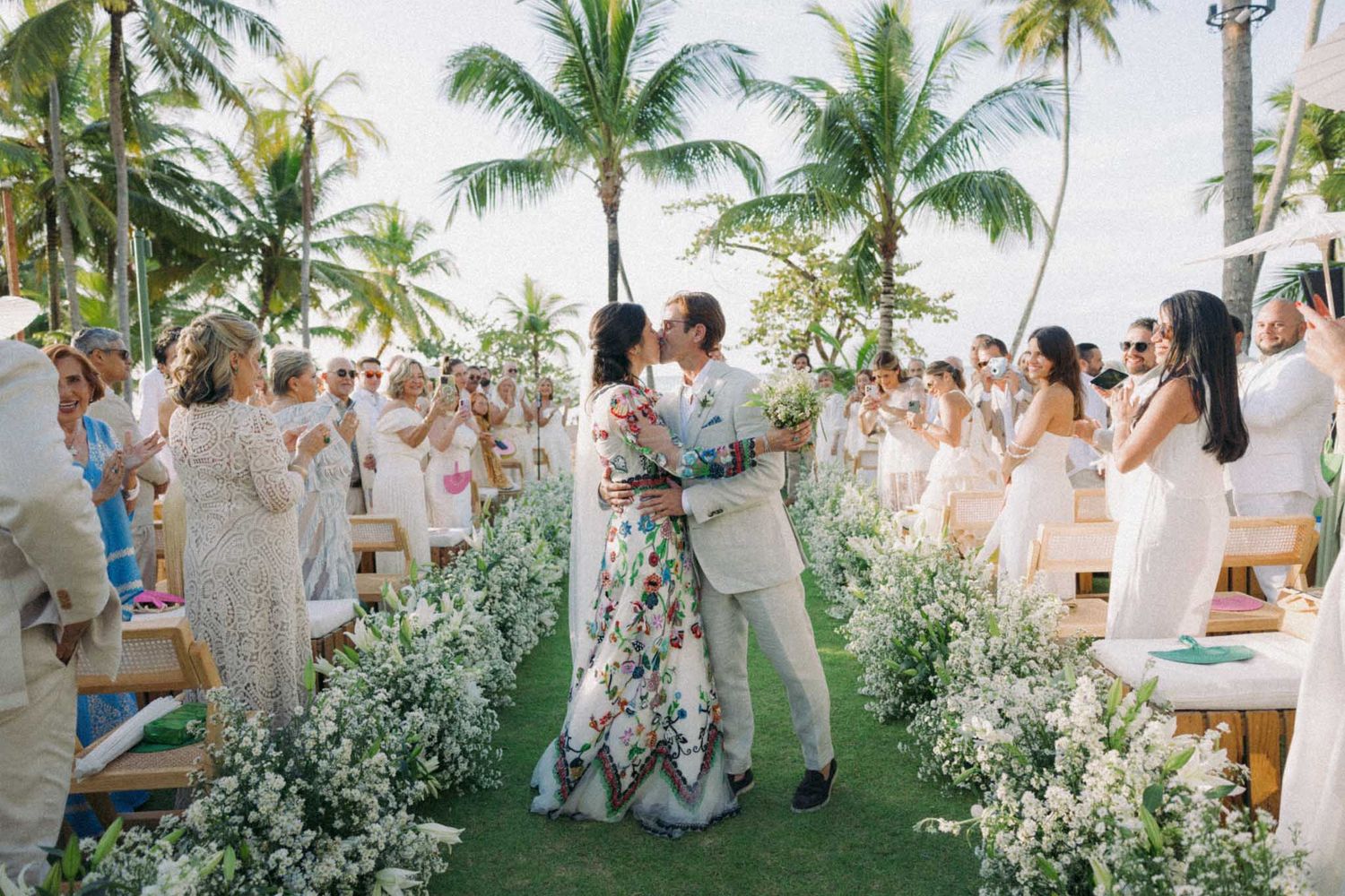 Tropical wedding ceremony setup with white chairs and floral arrangements under palm trees on a beachfront lawn.