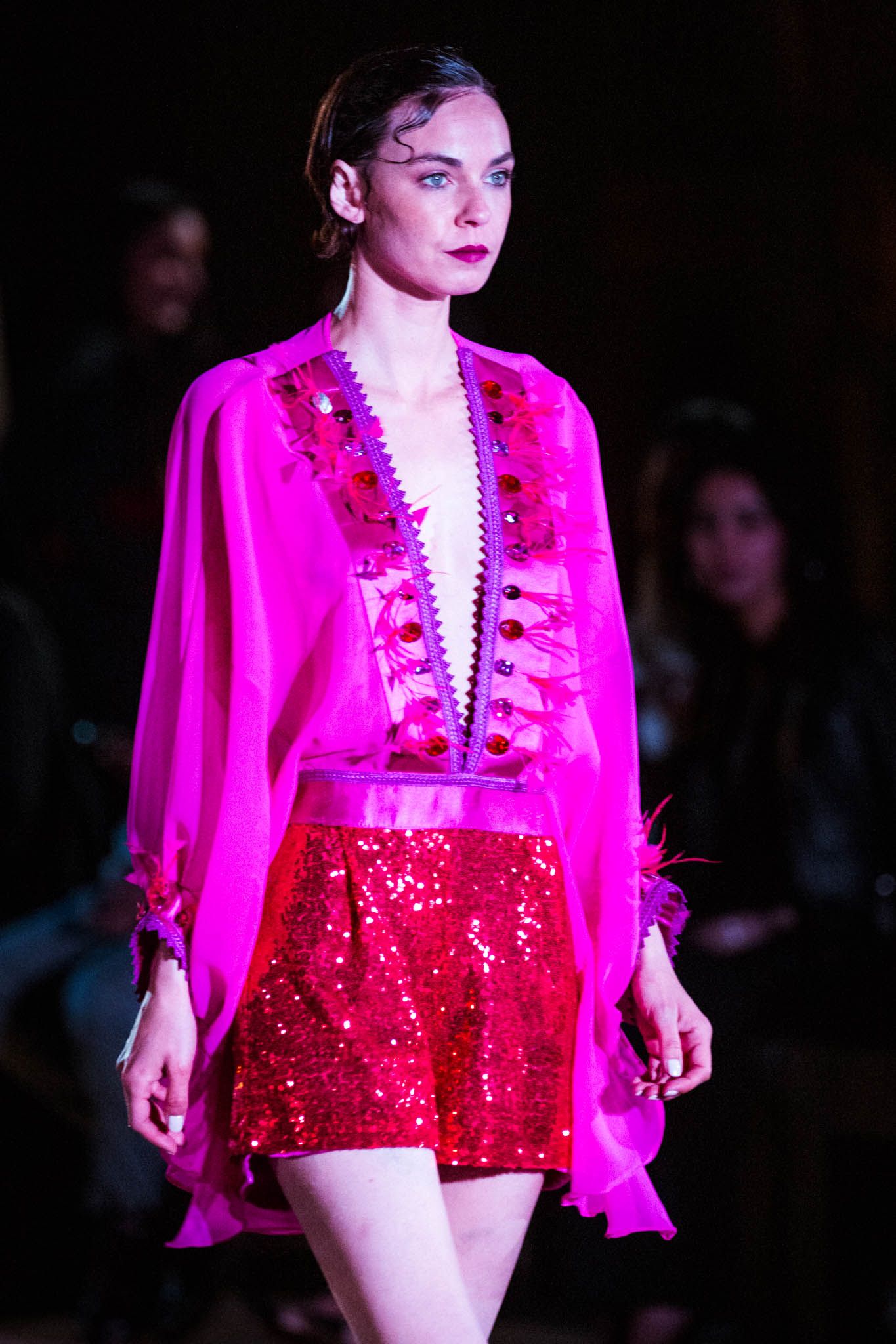 Model walks runway wearing bright fuchsia silk blouse with ruffled details and sparkly red mini skirt during fashion show.
