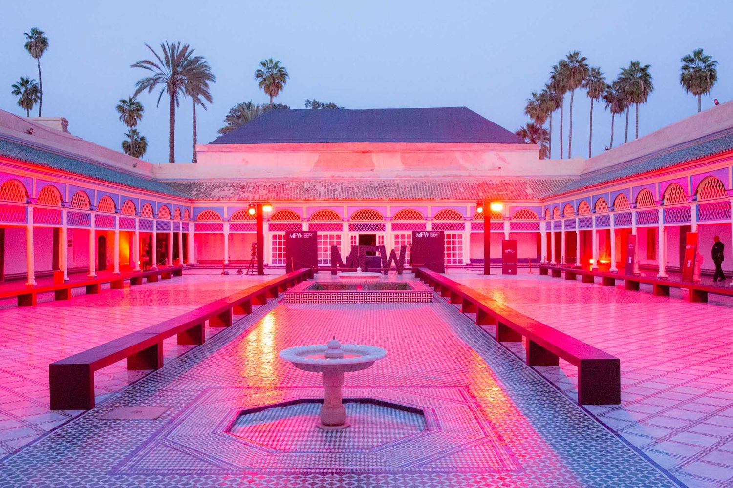 Moroccan courtyard illuminated in vibrant pink light with reflecting pool and palm trees at dusk.
