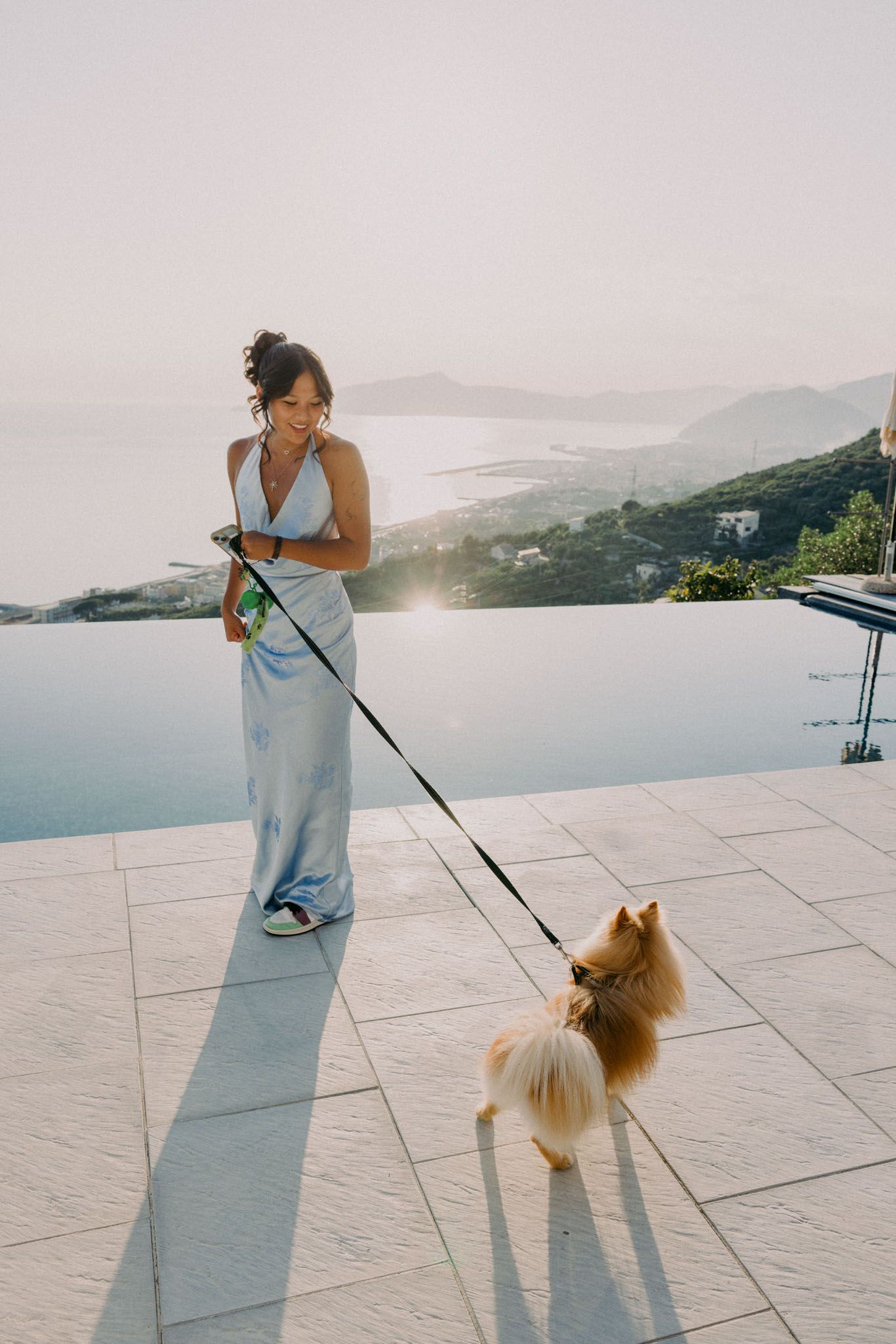 Person walking a fluffy Pomeranian dog on a scenic terrace overlooking mountains and water during sunset.