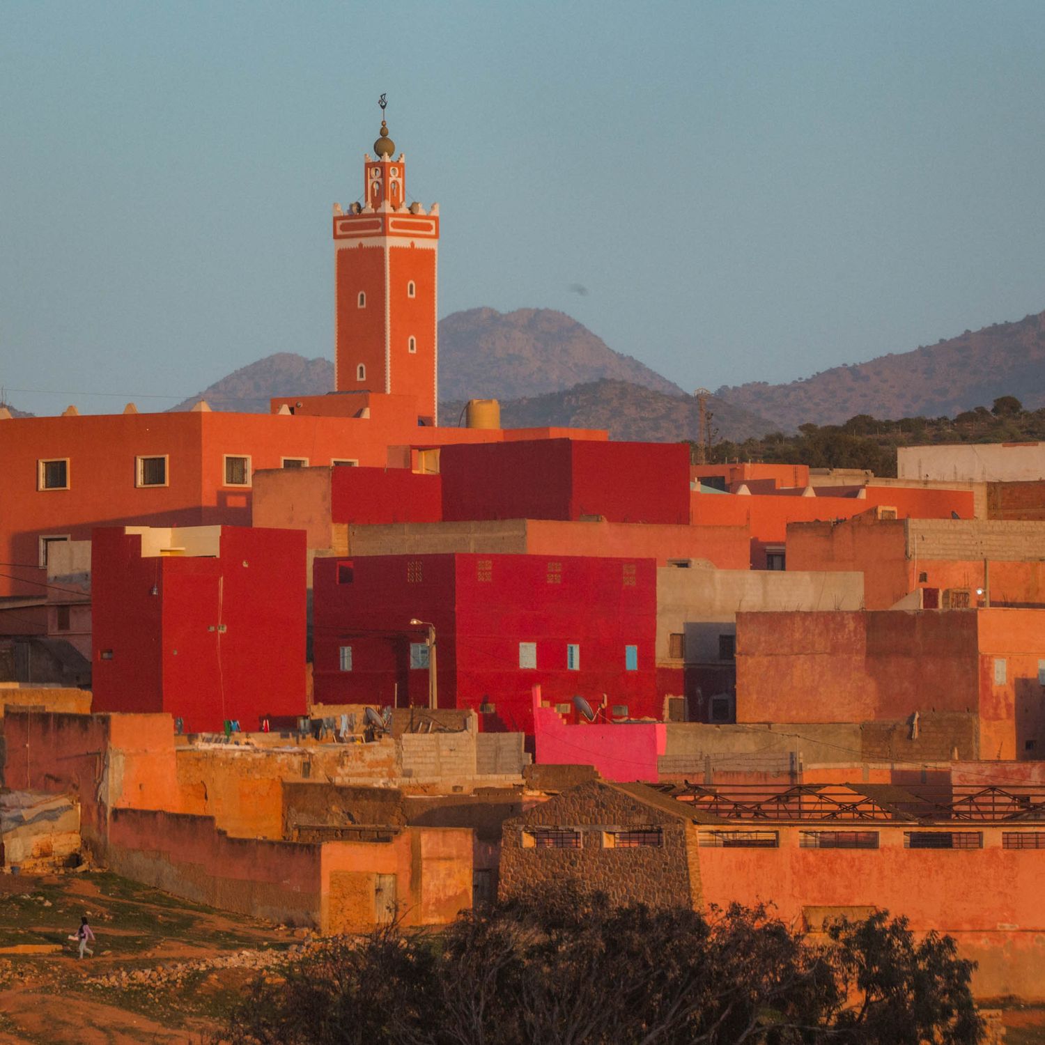 Vibrant orange and red buildings with towers dot the landscape against mountains in a traditional Moroccan village at sunset.