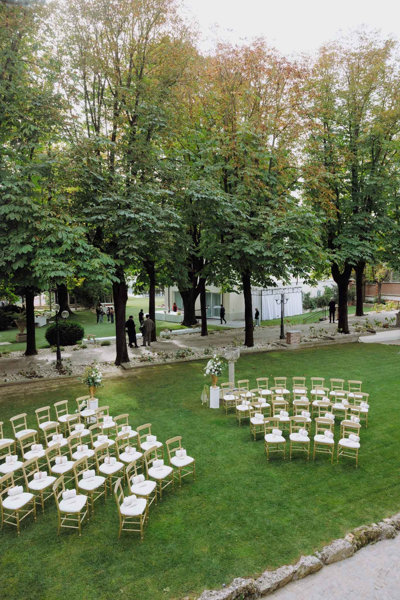 White folding chairs arranged in rows on a green lawn for an outdoor ceremony with tall trees in the background.