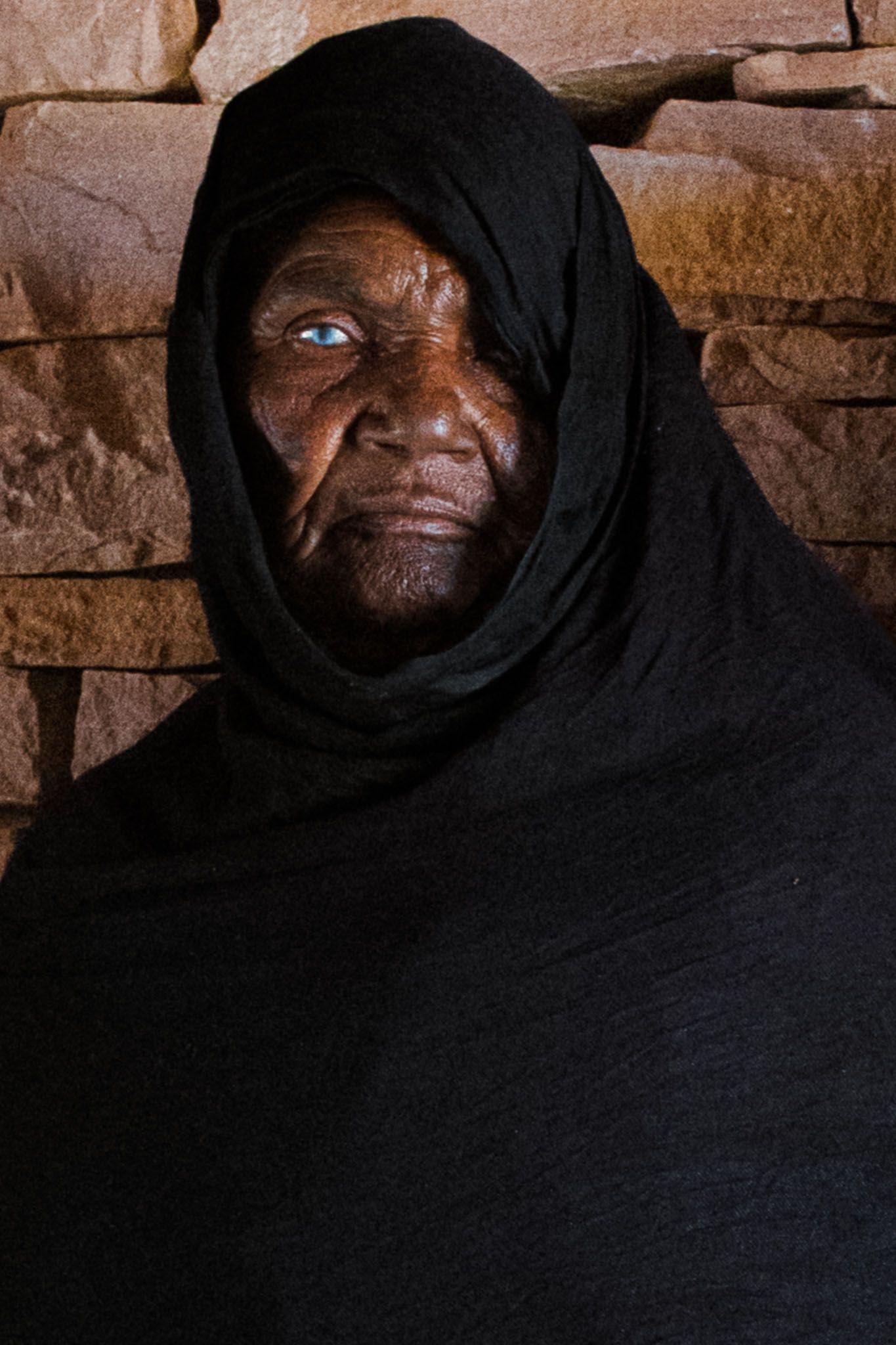 Close-up portrait in dramatic lighting showing traditional black headwear against stone wall.