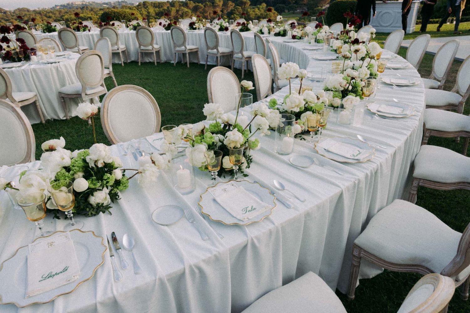Elegant outdoor wedding reception setup with white tables, chairs, and floral centerpieces arranged on a manicured lawn.