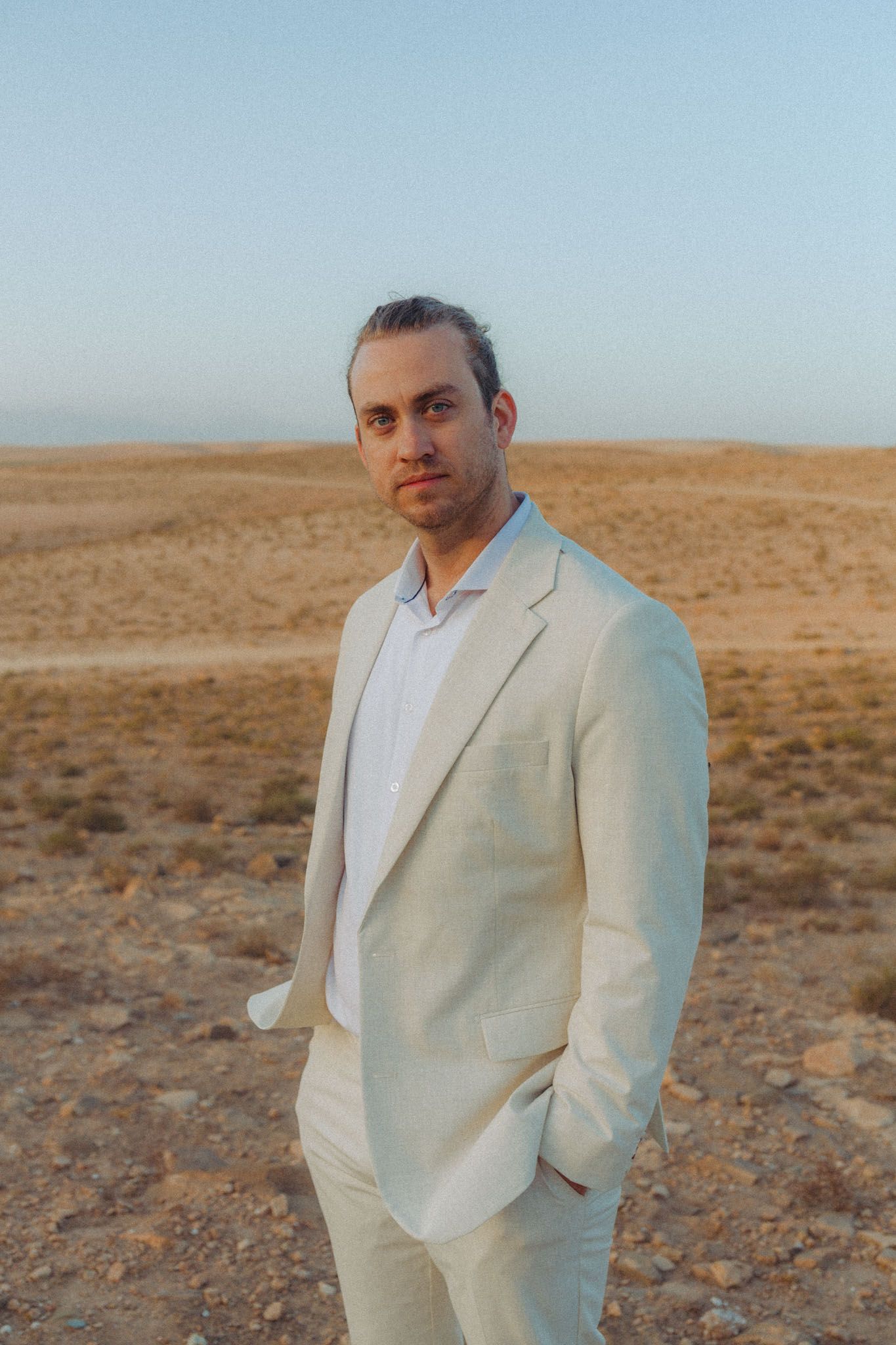 A person in a light-colored suit stands in a desert landscape against a barren, rocky terrain.