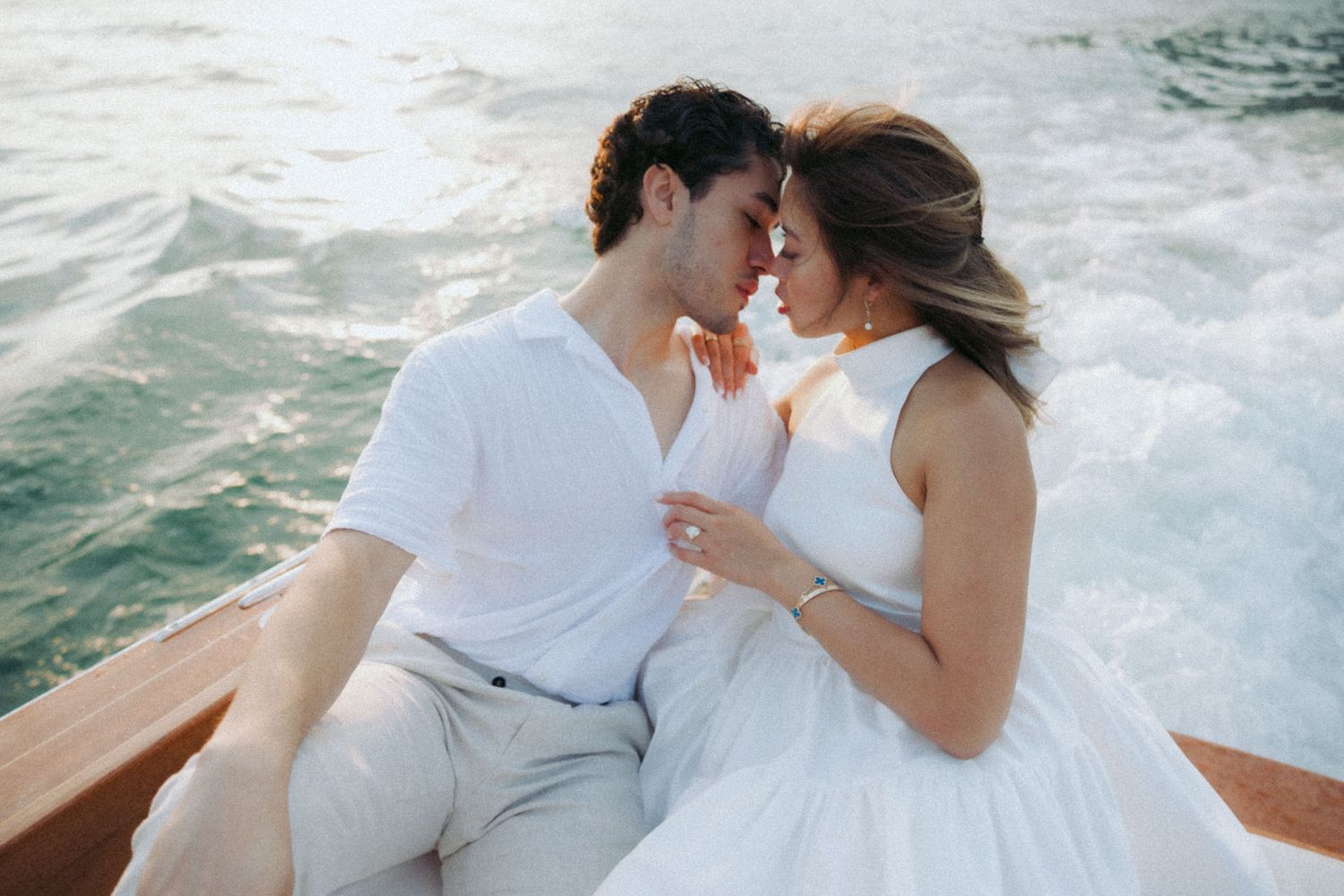 Romantic couple in white outfits sharing intimate moments on a boat against splashing water background.