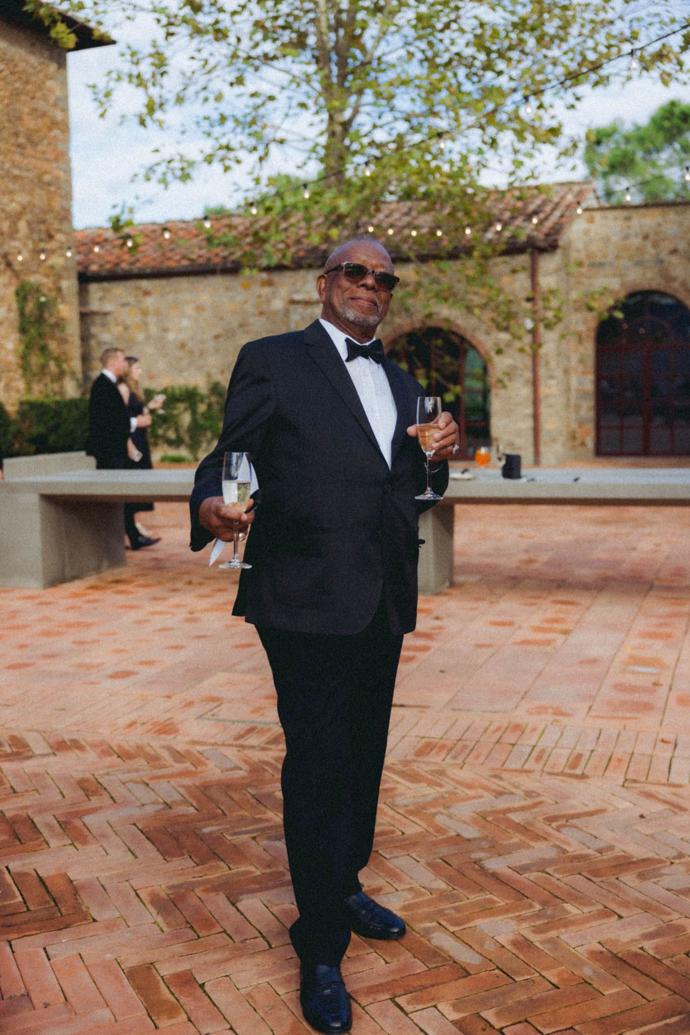 A person in a black tuxedo and sunglasses walks across a brick courtyard with a wine glass in hand.