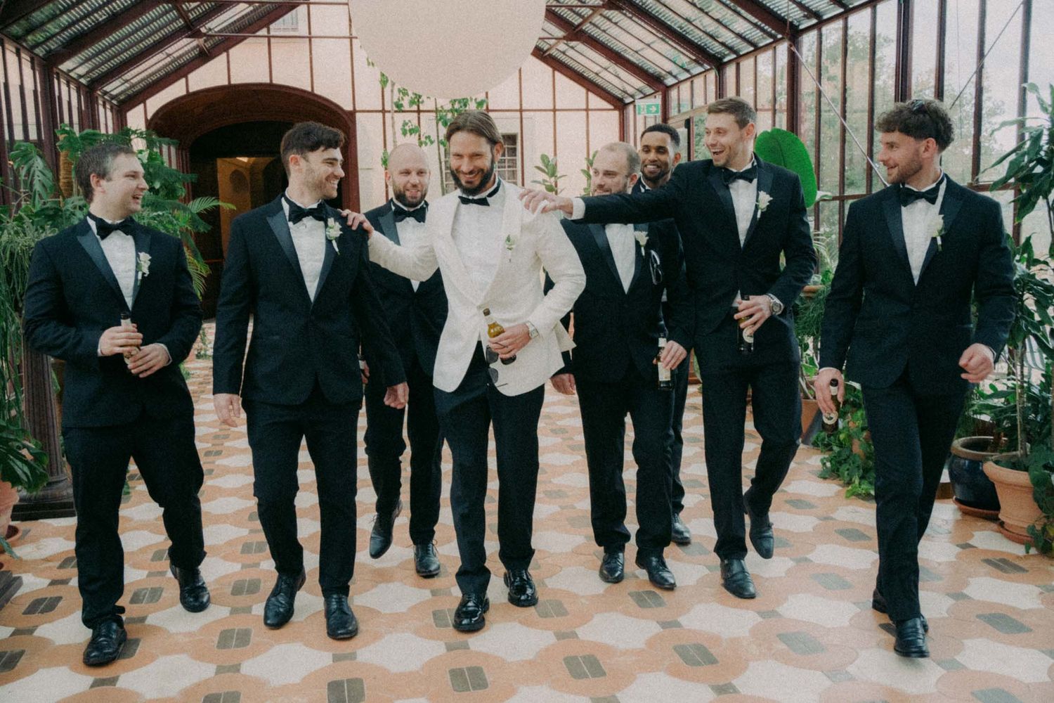 Groomsmen in black tuxedos and bowties walk in line through a greenhouse conservatory.