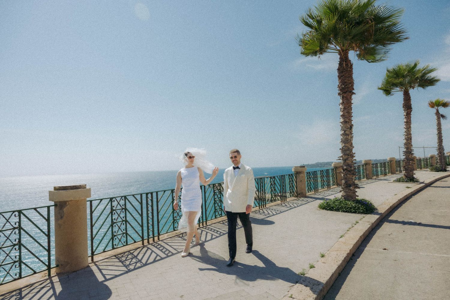 Scenic beachfront walkway with palm trees and turquoise railings overlooking white sandy shores and blue ocean waters.