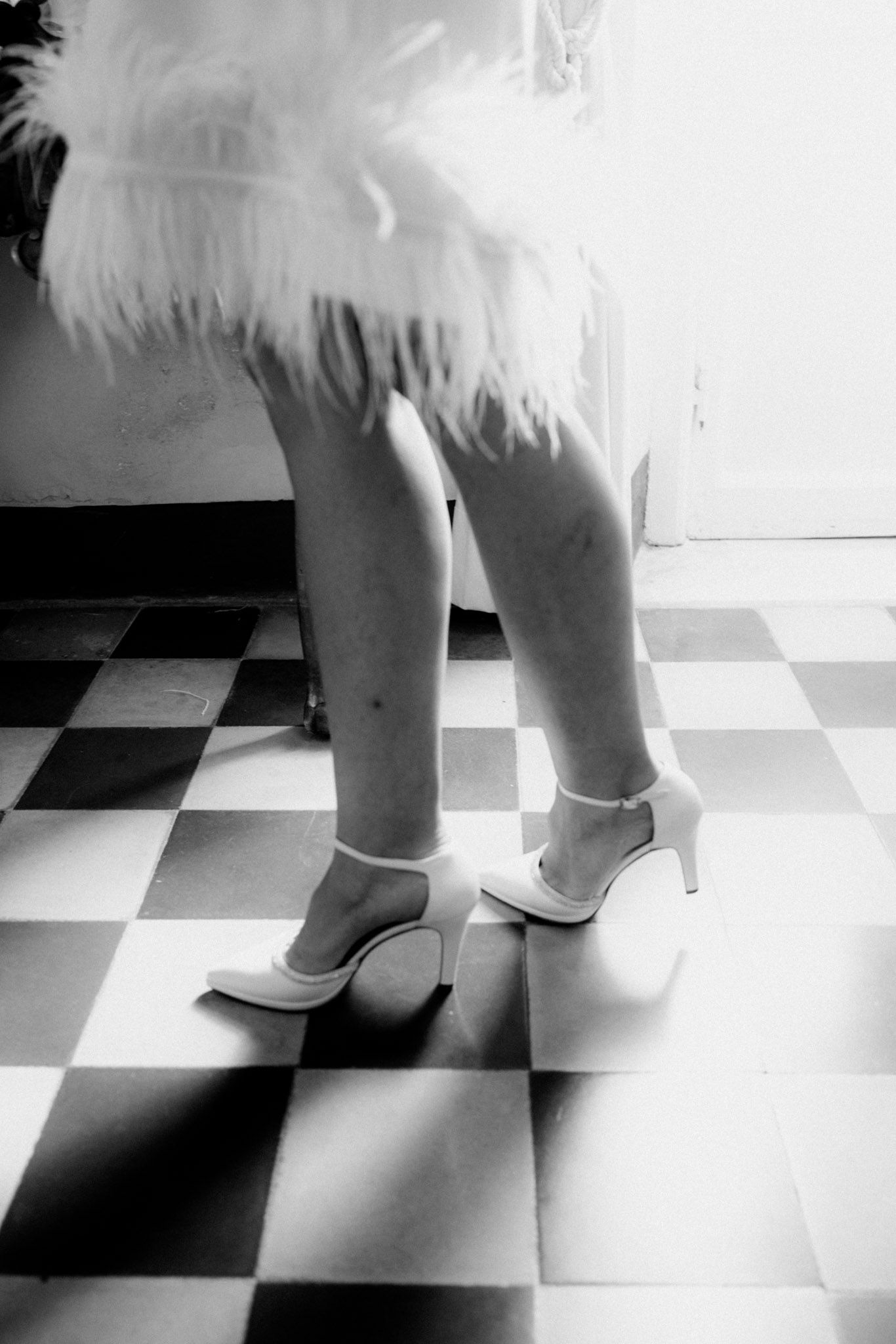 Black and white photo of legs in white high heels standing on a checkered floor with feathers visible above.