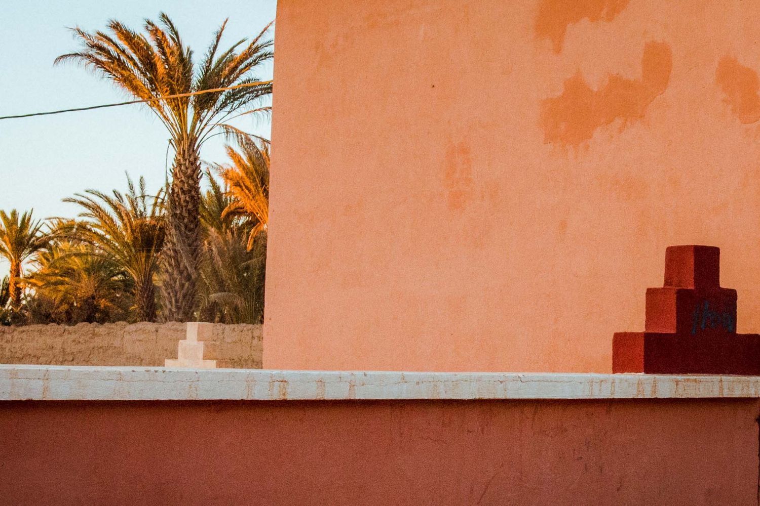 Orange stucco wall with palm trees and architectural details casting shadows in warm sunlight.