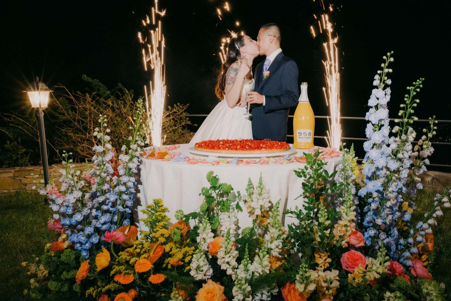 A couple shares a romantic moment behind their wedding cake surrounded by colorful flowers and sparkler fountains at night.