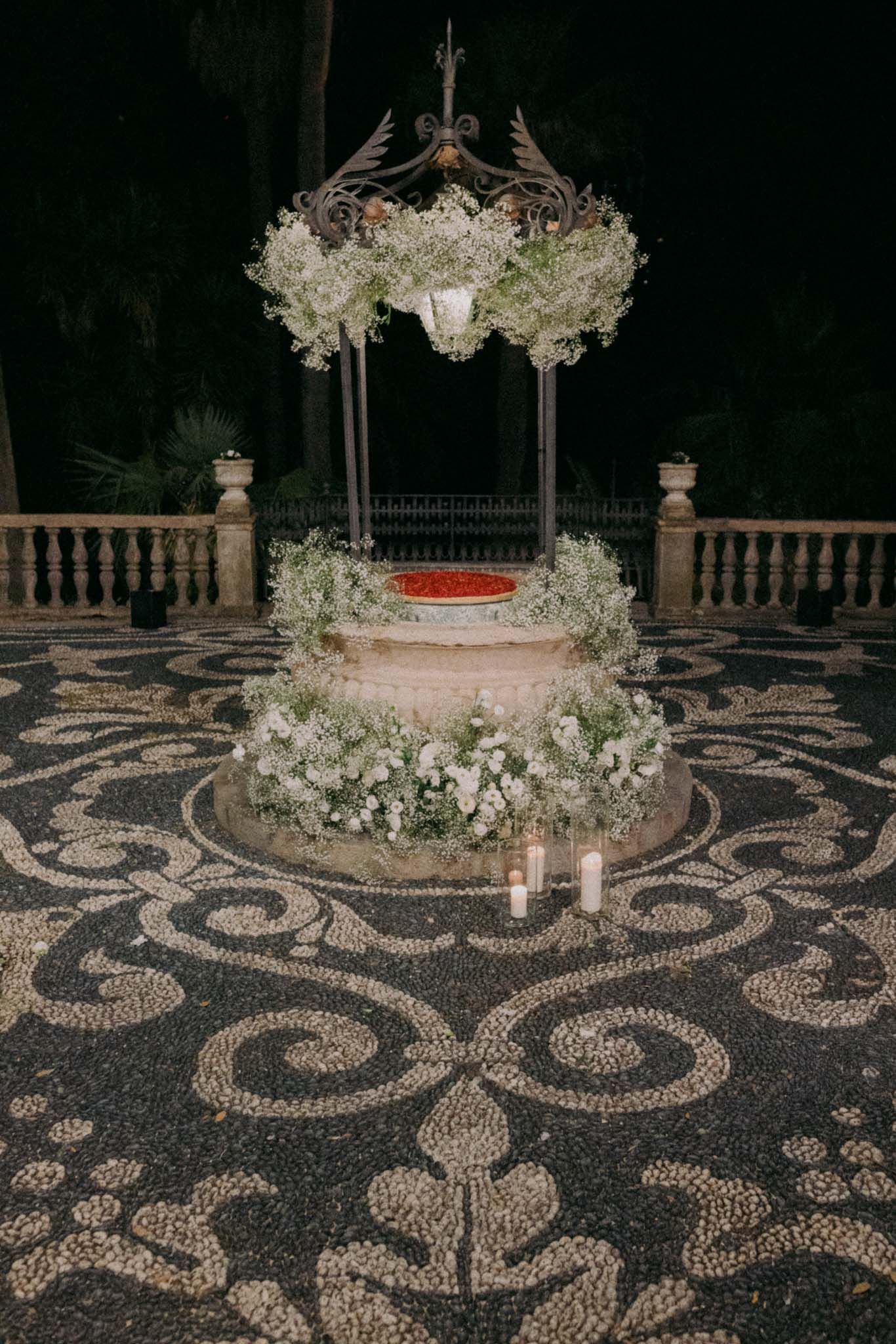 Elegant wedding cake display on ornate mosaic floor with white floral decorations and canopy at night.