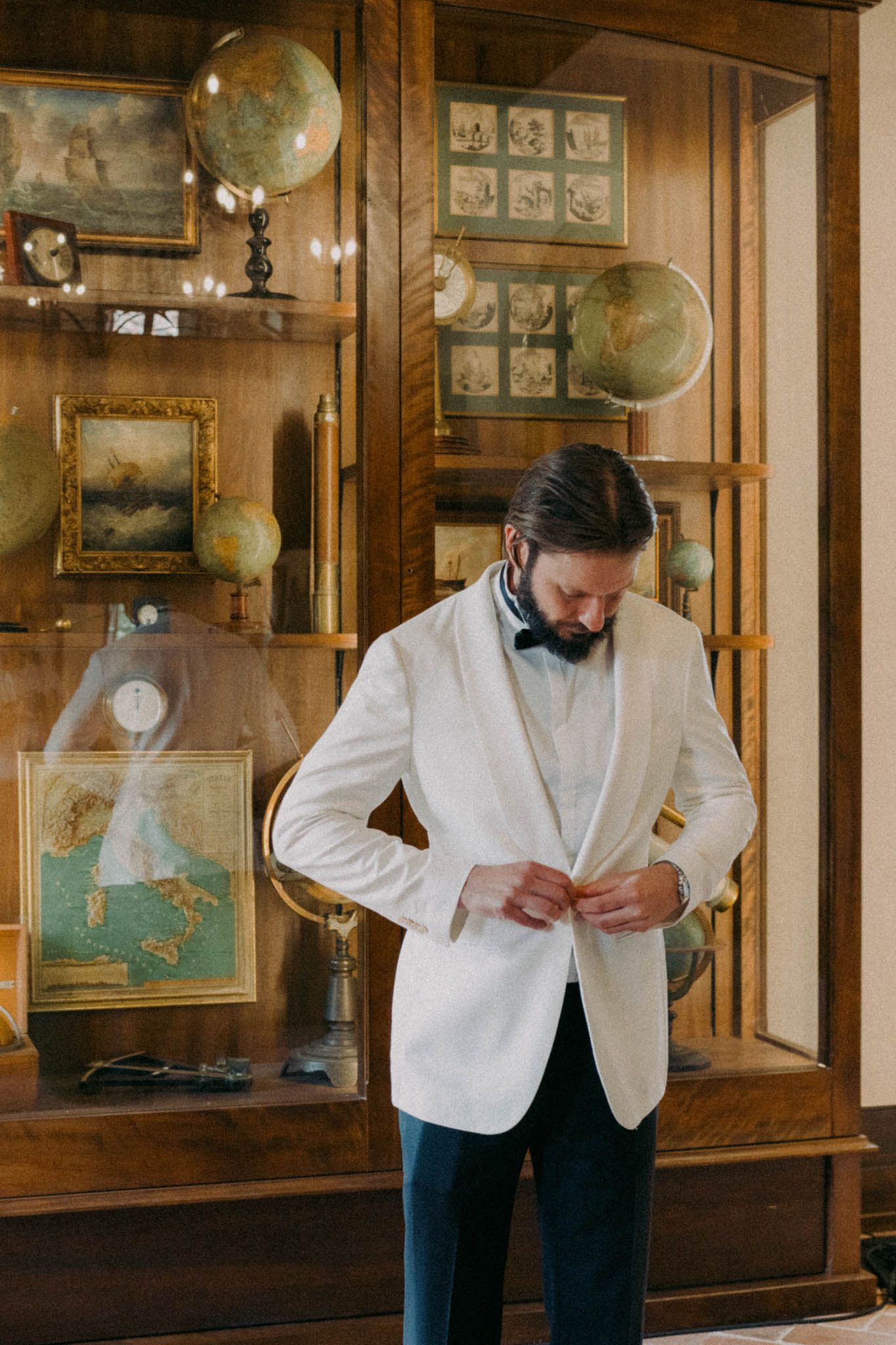 A person in a white dinner jacket and black pants adjusts their attire in an elegant wood-paneled room with antique decor.