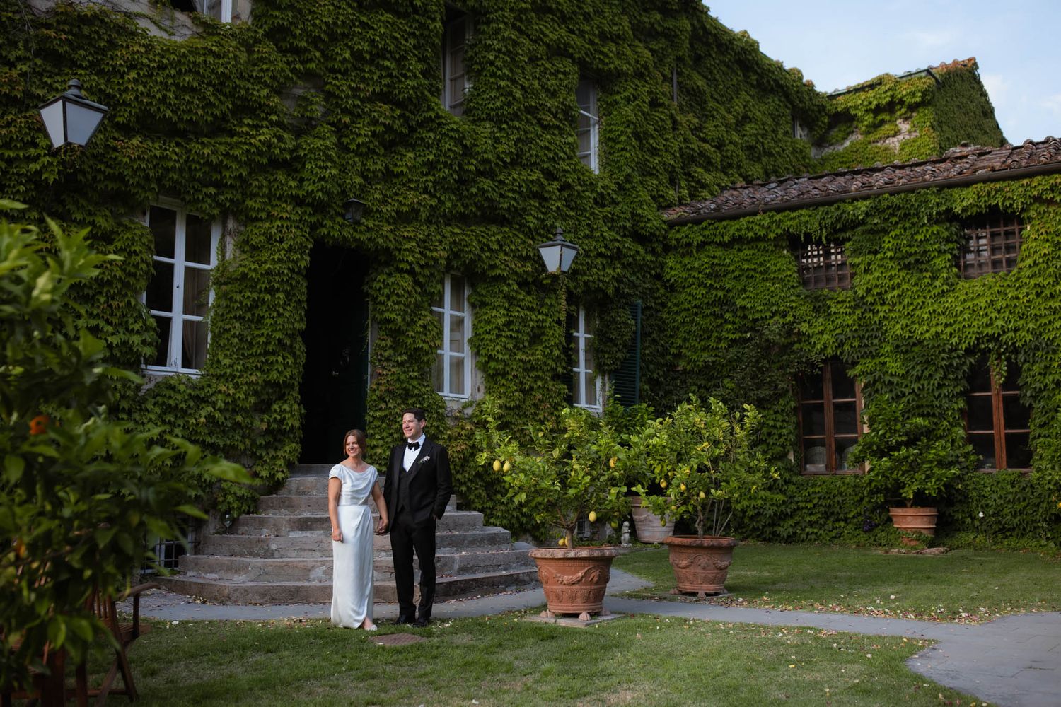 Elegant figures walk through a lush garden path lined with ivy-covered walls and vintage lampposts.