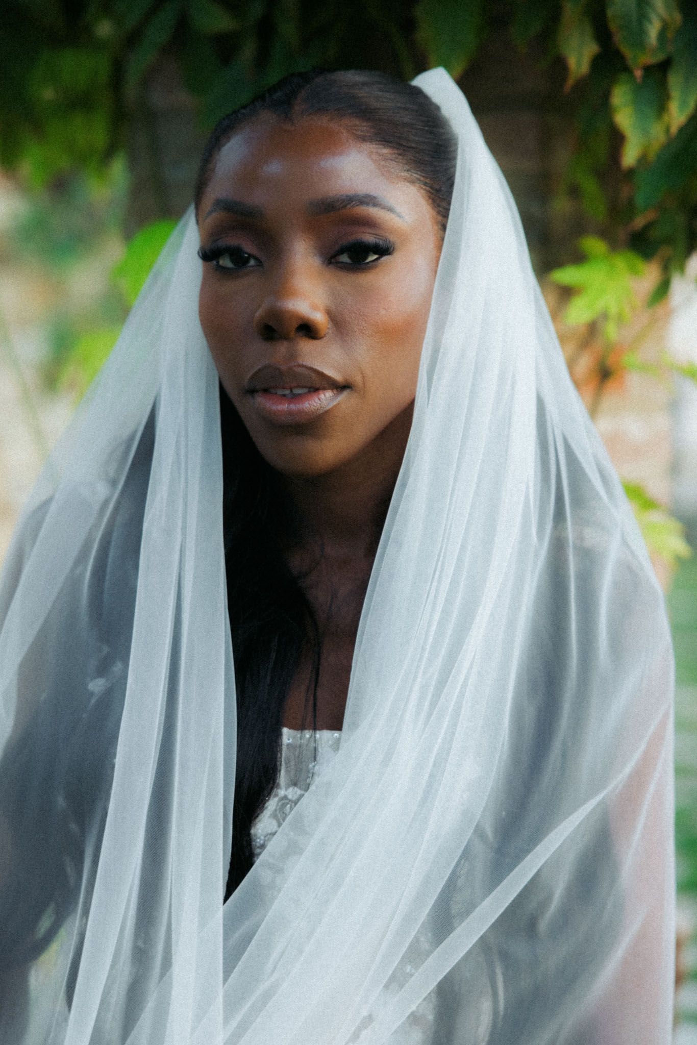 A bride wearing a flowing white veil poses in a natural outdoor setting with soft lighting and greenery in the background.