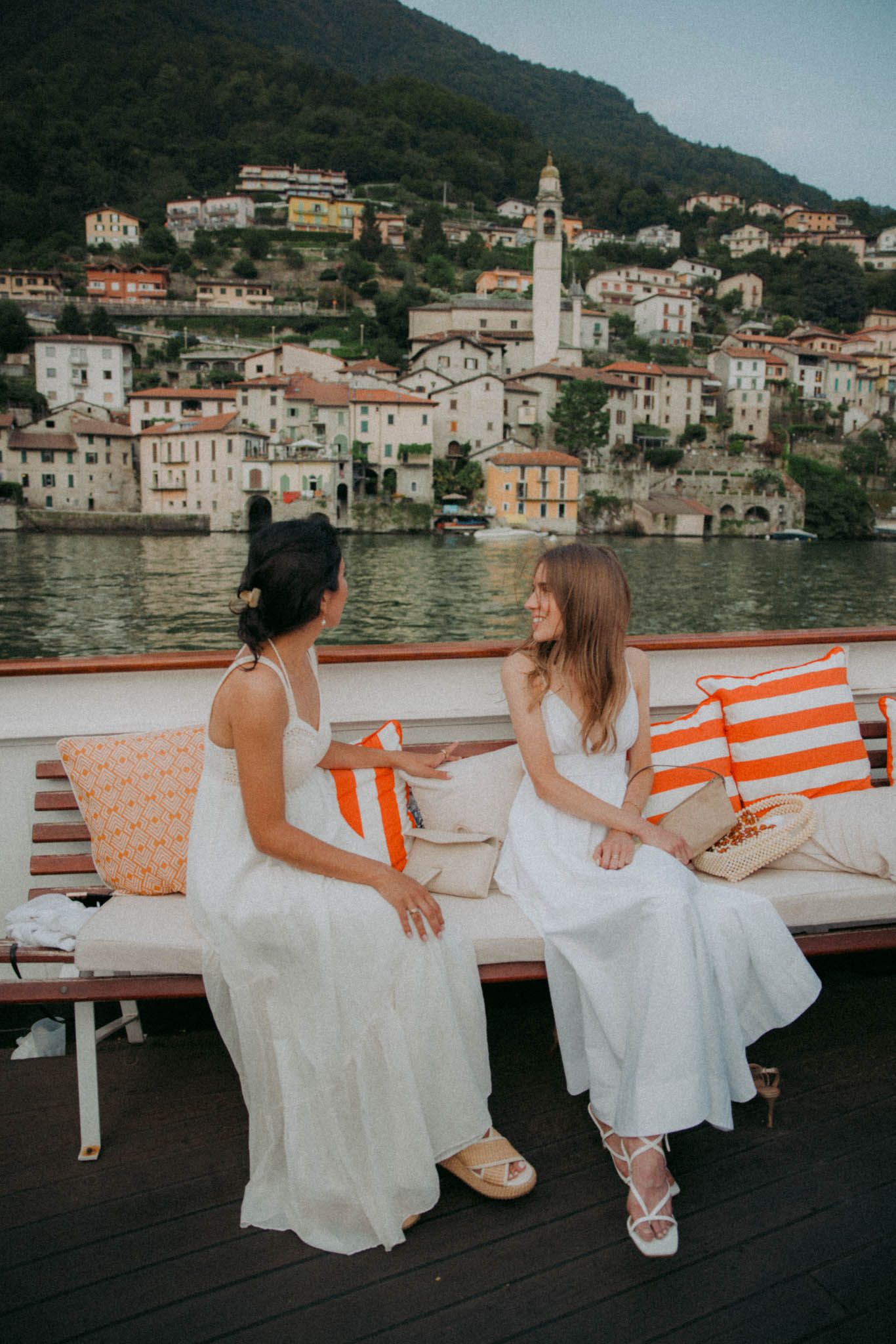 Two people in white dresses sitting on a boat deck overlooking a scenic Italian coastal village with historic buildings.