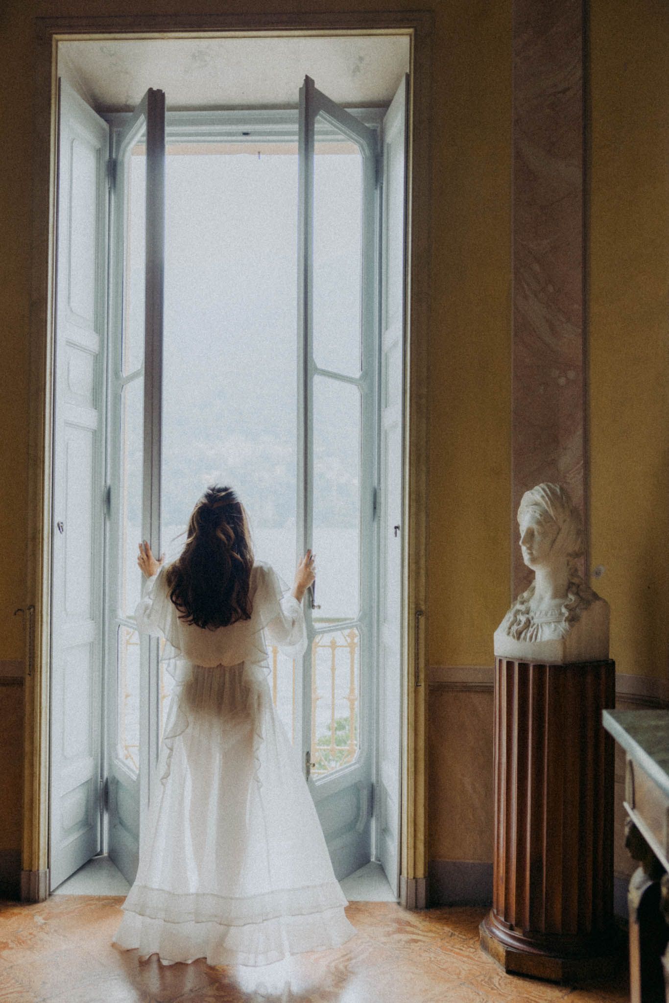 A ghostly figure in white flowing dress stands in front of multiple windows in an elegant interior space.